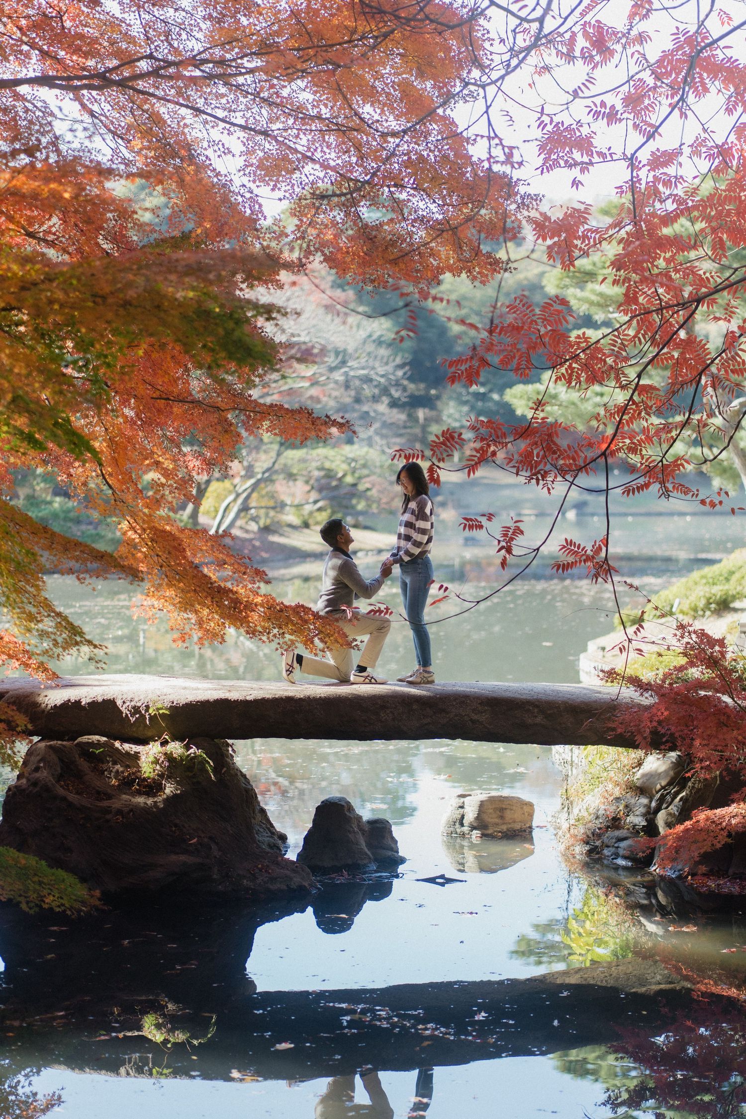 Couple on stone bridge amid vibrant autumn maple trees in a Japanese garden, with serene pond reflection below.