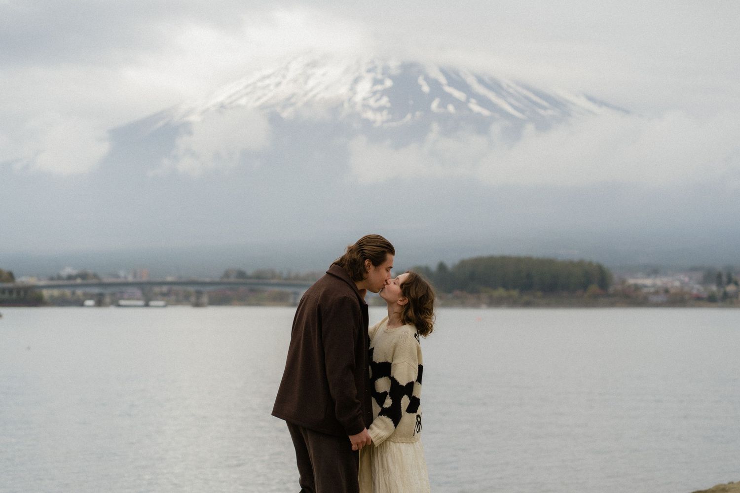Couple sharing a romantic kiss by a lake with snow-capped Mount Fuji in the background on a cloudy day.