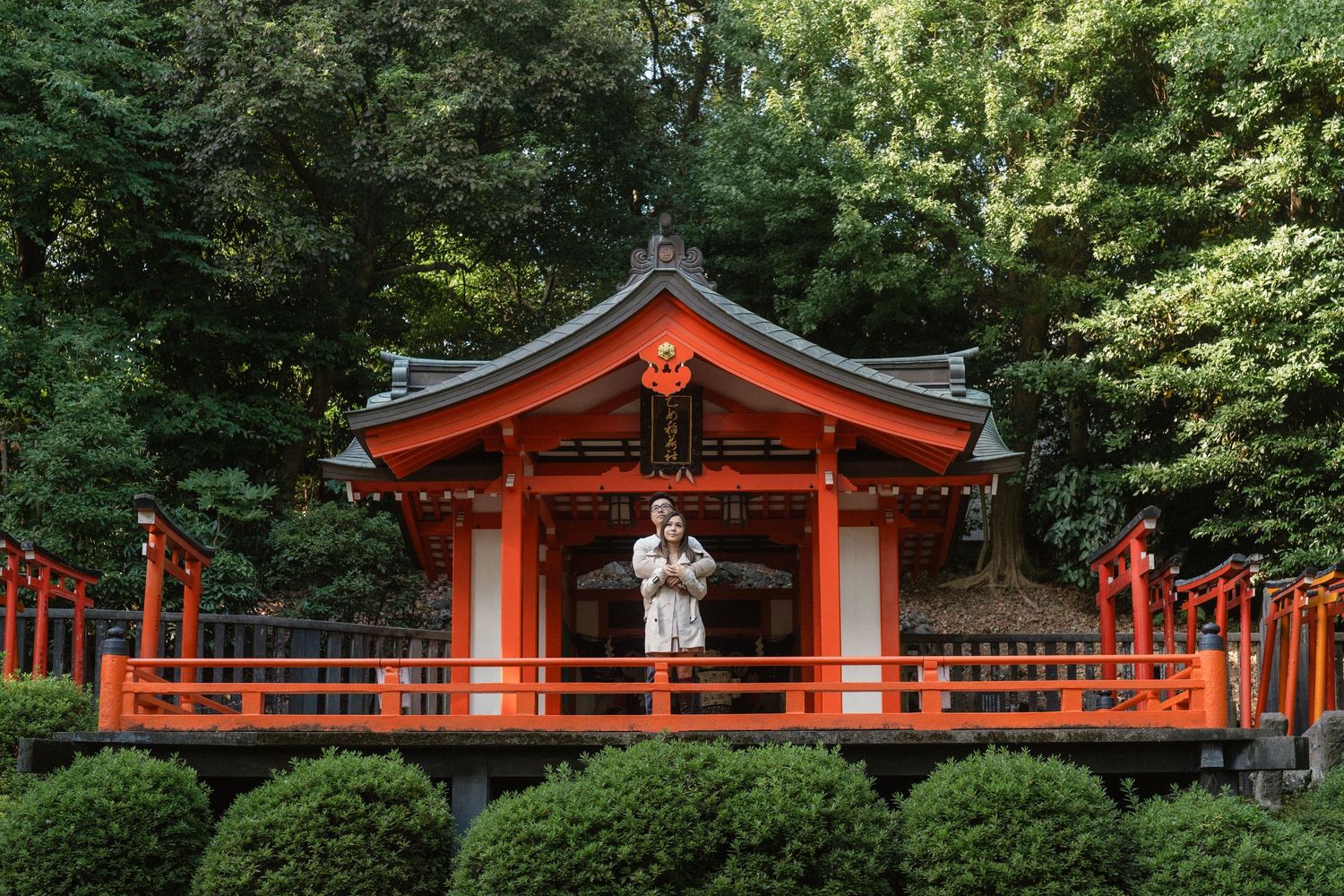 Woman in white kimono standing on red bridge at traditional Japanese shrine surrounded by lush green trees.