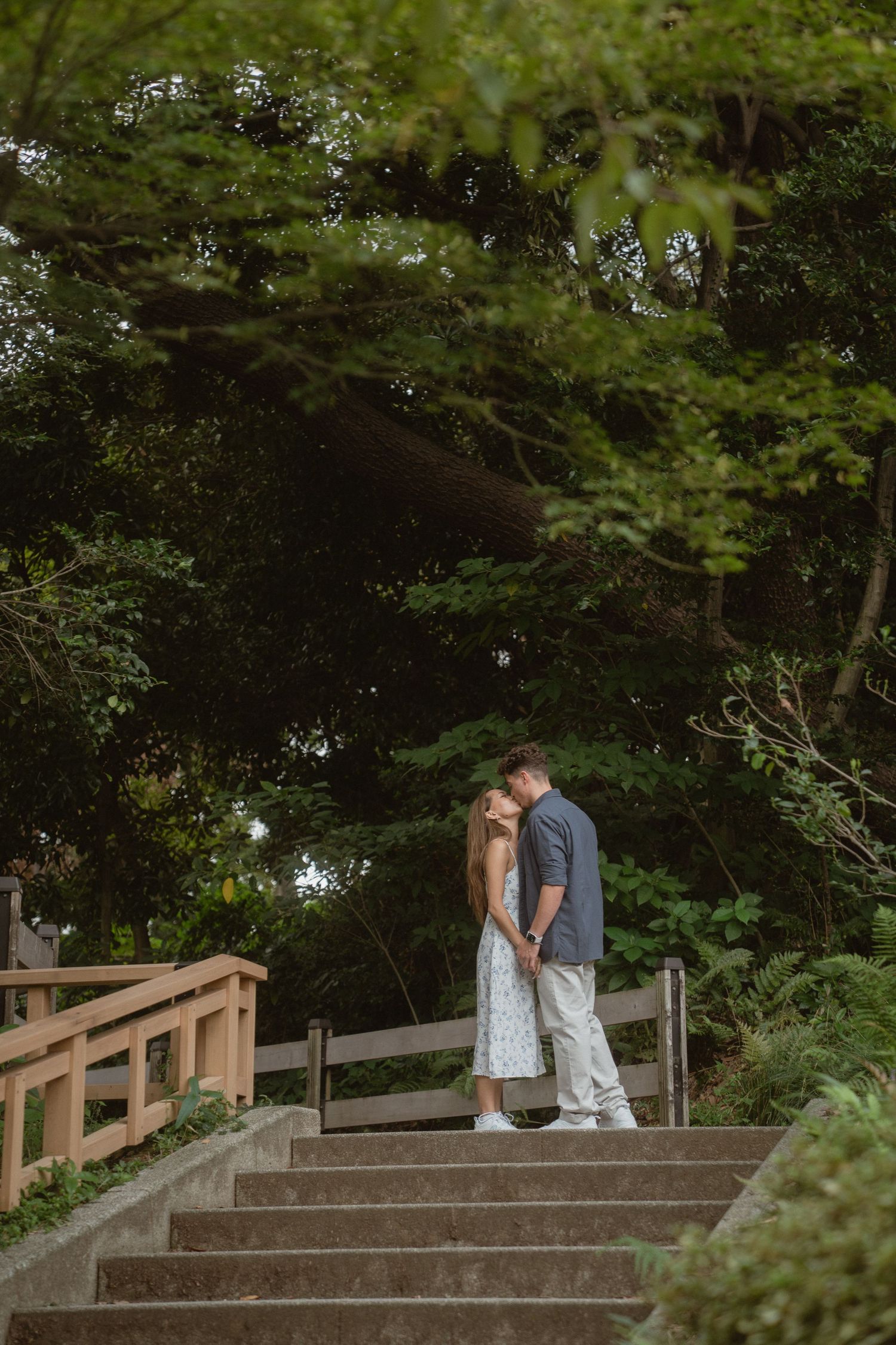Couple sharing a romantic kiss on garden steps surrounded by lush green trees in a serene outdoor setting.