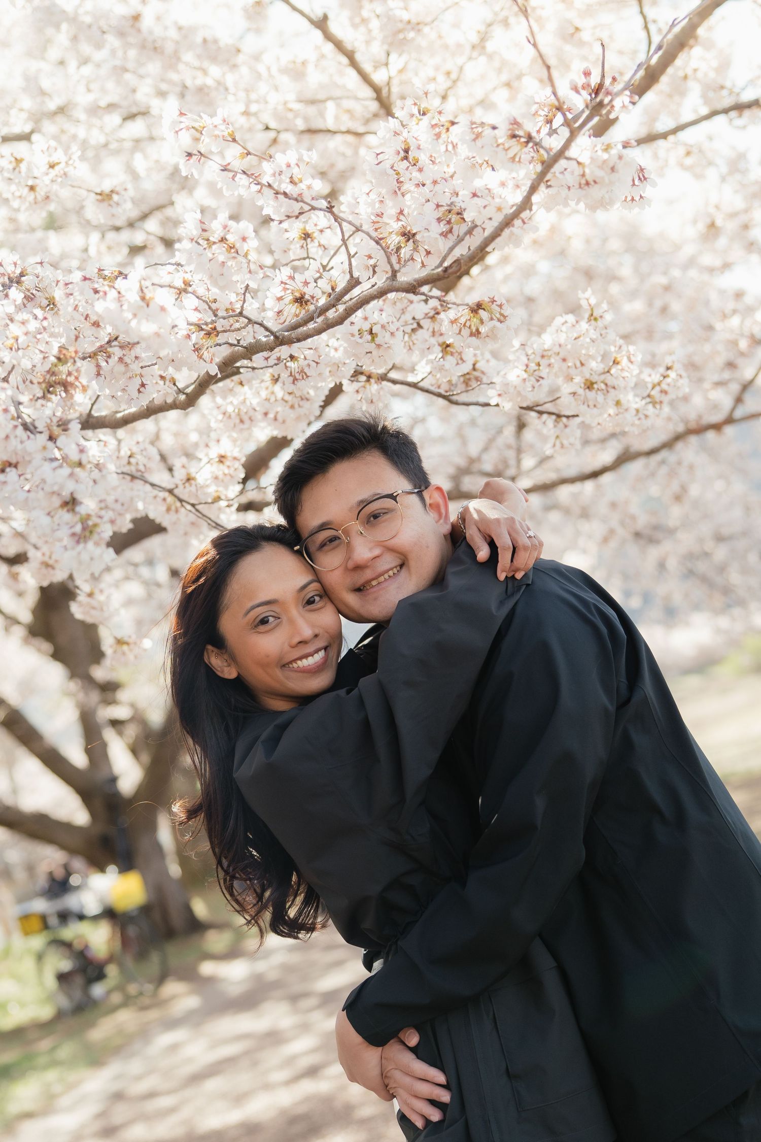 Happy couple embracing under blooming cherry blossom trees in spring, smiling at camera outdoors.