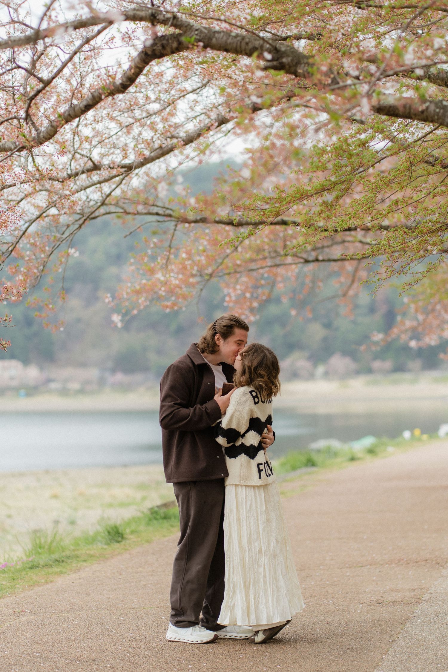 Couple sharing a romantic kiss under blooming cherry blossom trees near a tranquil lake in spring.