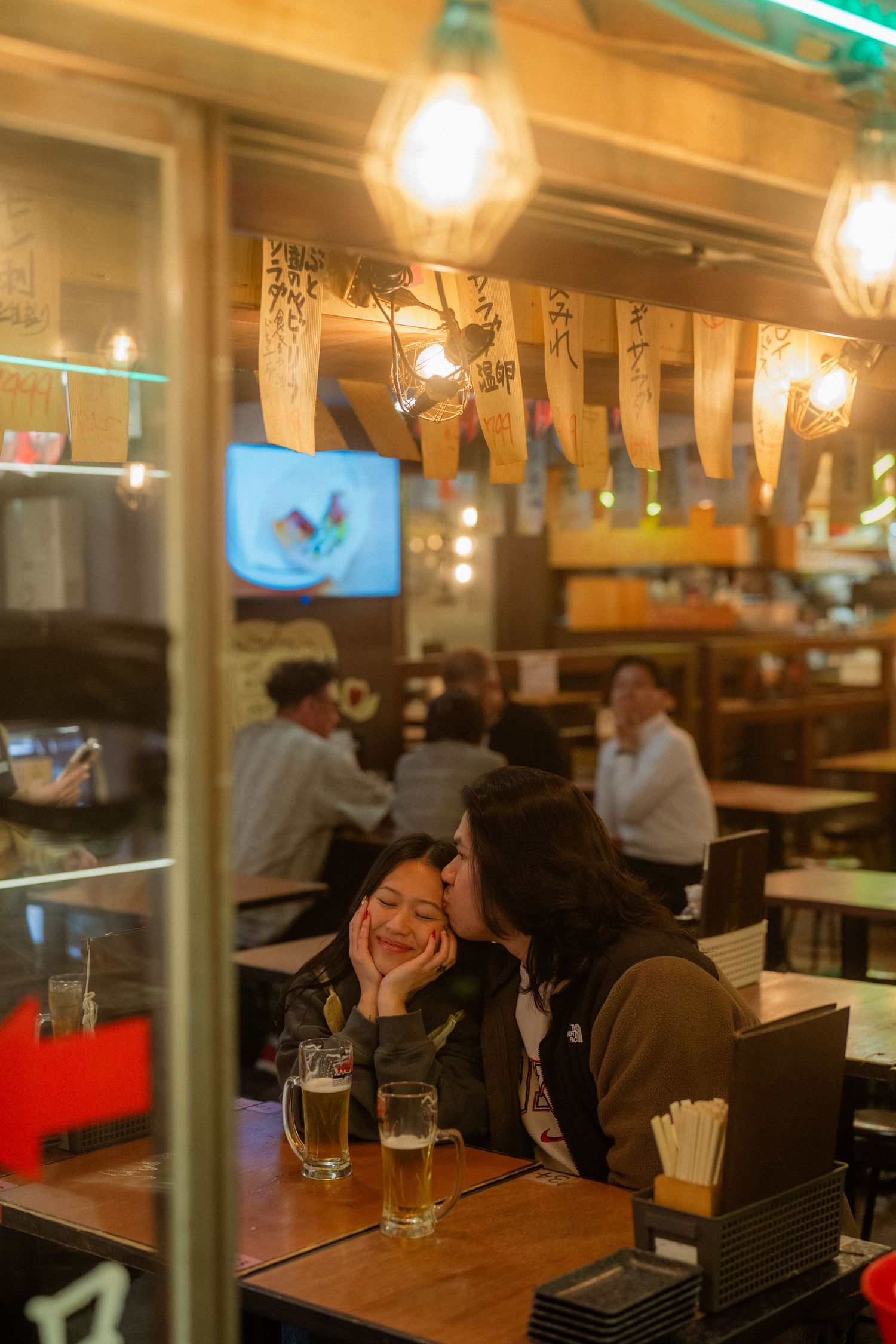 Two women share a kiss at a cozy Japanese izakaya bar with beer glasses and hanging banners.