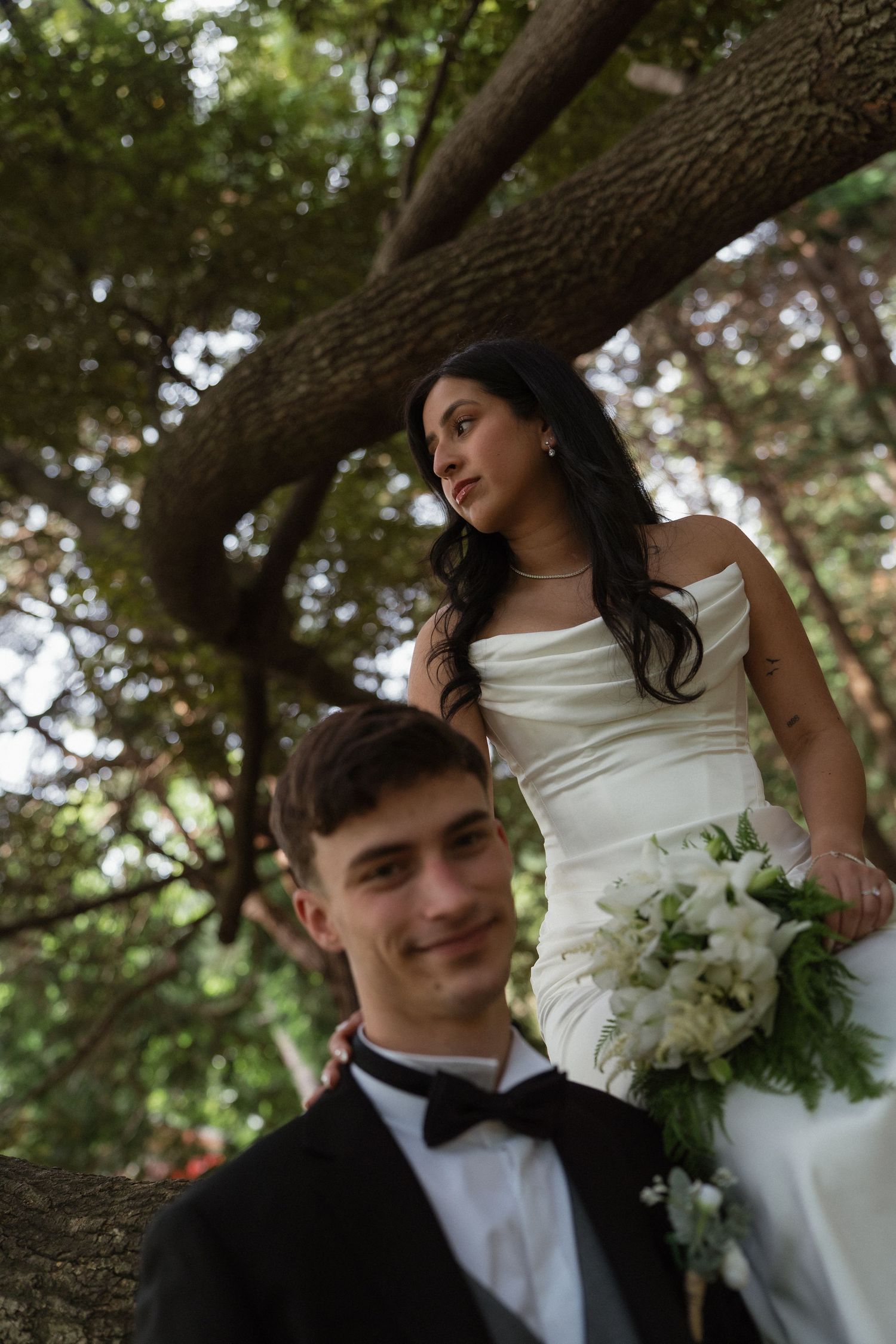 Bride in white strapless gown holding bouquet leans on tree while groom in black tuxedo smiles in foreground.