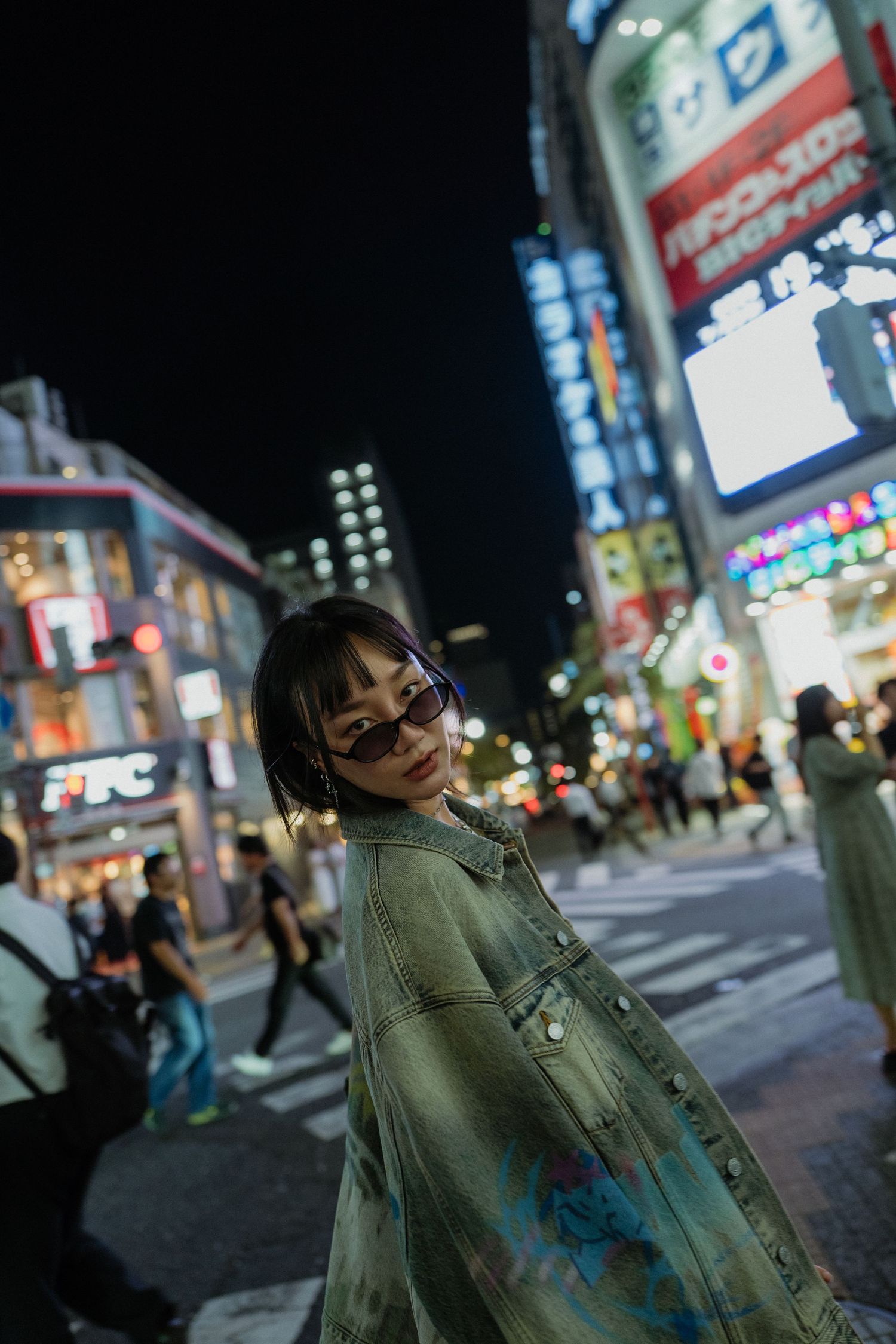 A young woman in a denim jacket and sunglasses poses at a busy Tokyo crossing at night with neon signs.
