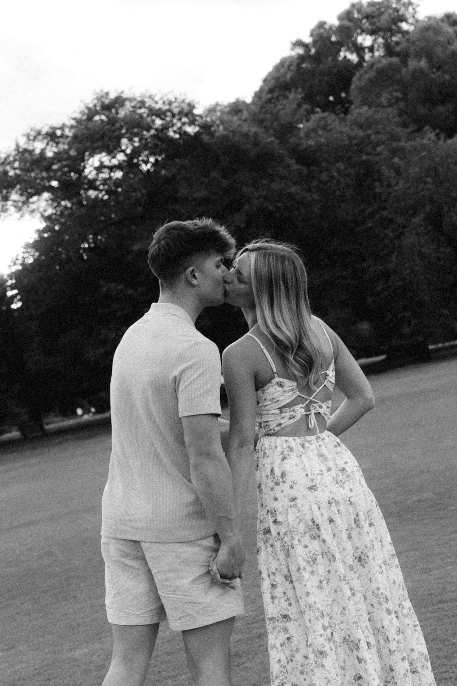 Black and white photo of a couple kissing while holding hands in a park with trees in the background.