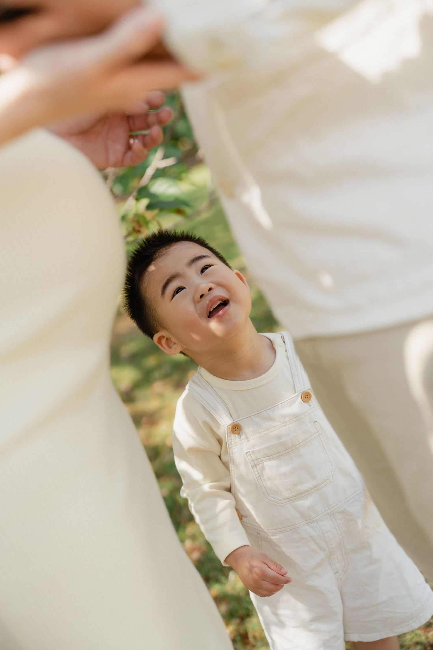 A joyful toddler in cream overalls laughs up at parents in a sunlit outdoor setting.