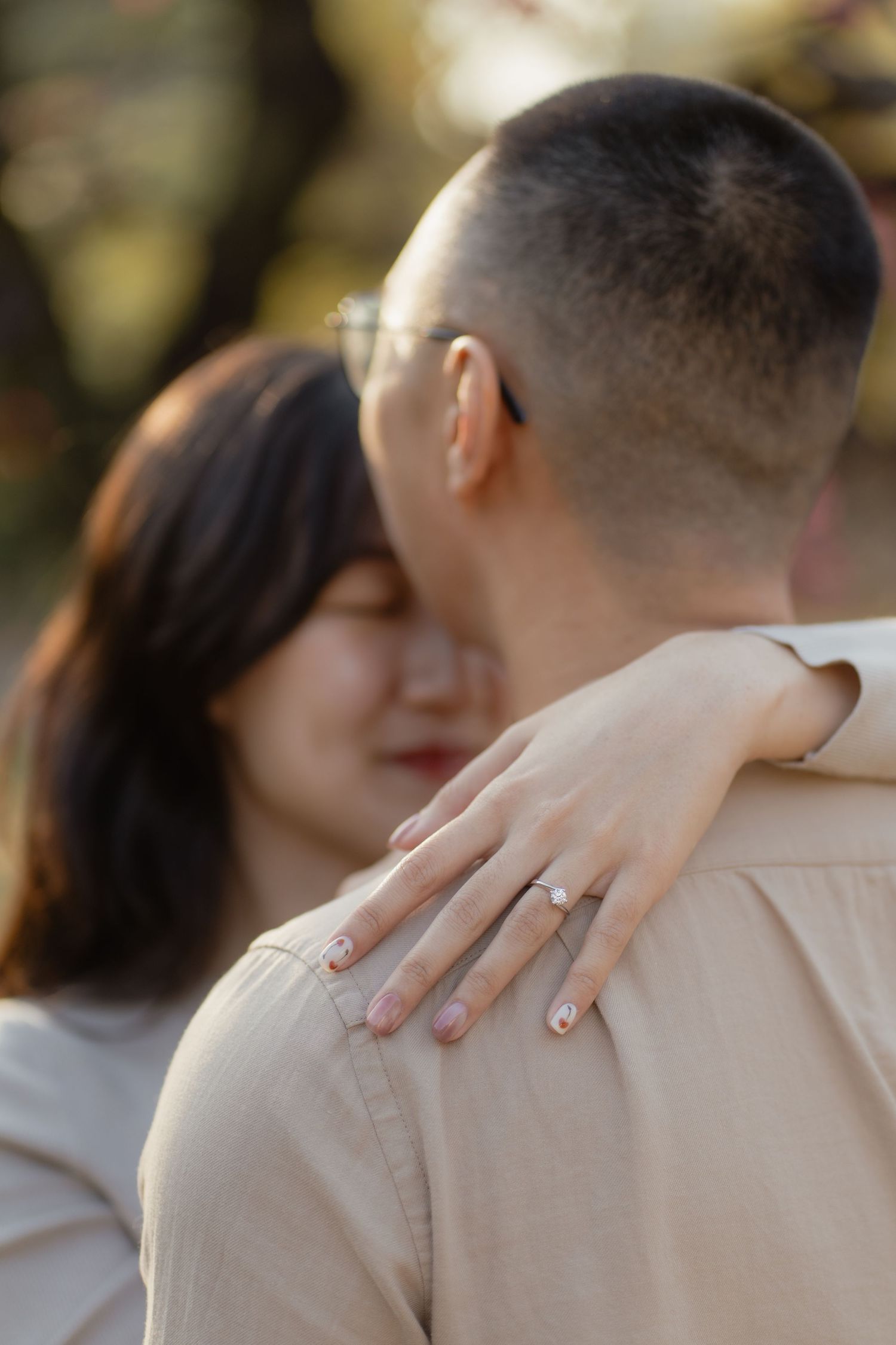 Couple embracing outdoors, woman resting head on man's shoulder showing engagement ring, soft bokeh background.
