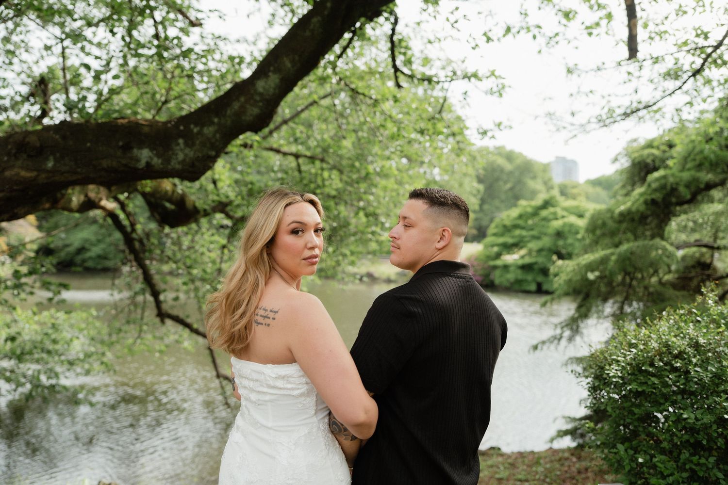 Bride in white strapless gown and groom in black suit pose near a serene pond surrounded by lush green trees.