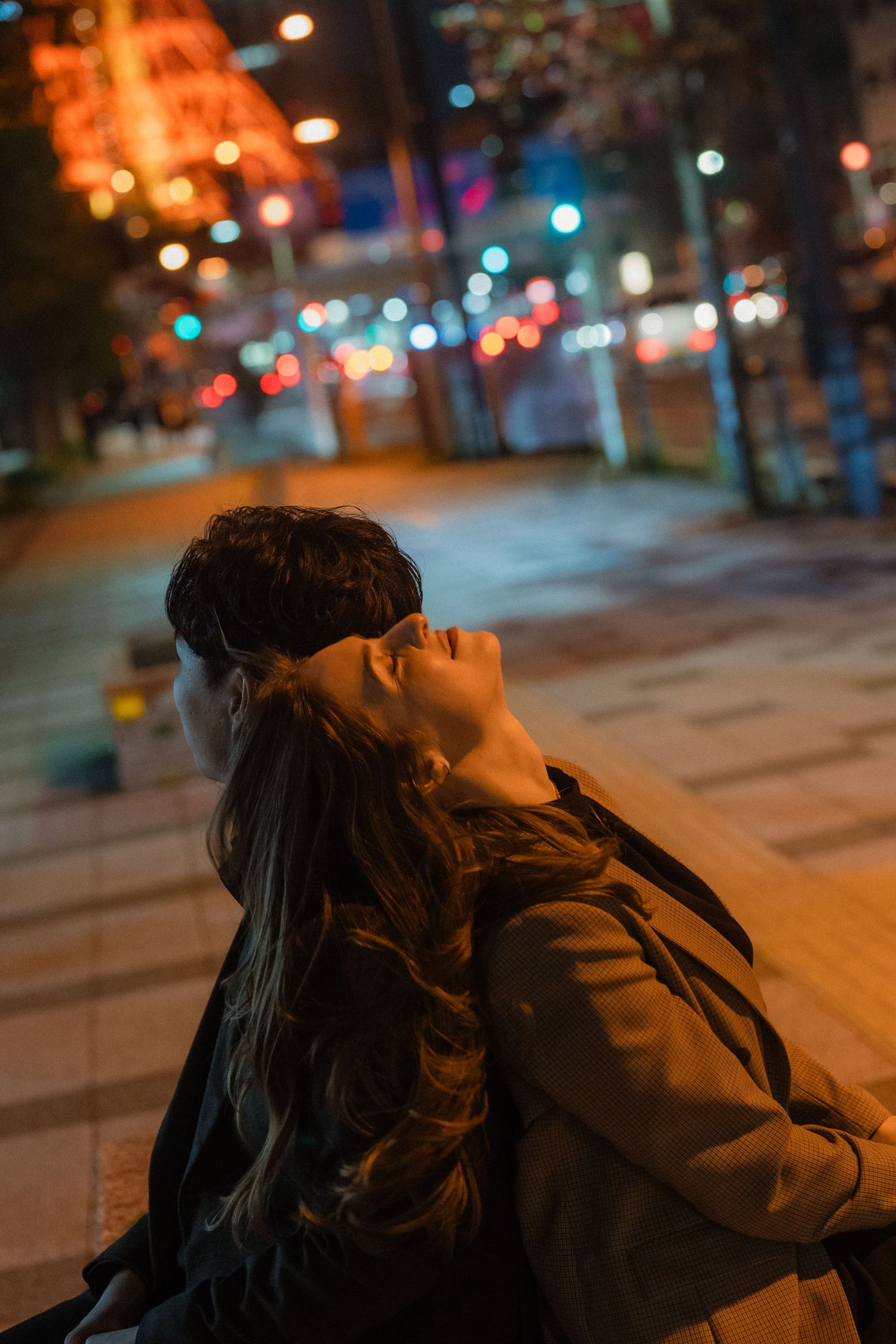 Couple embracing on a city sidewalk at night with colorful bokeh lights in the background.