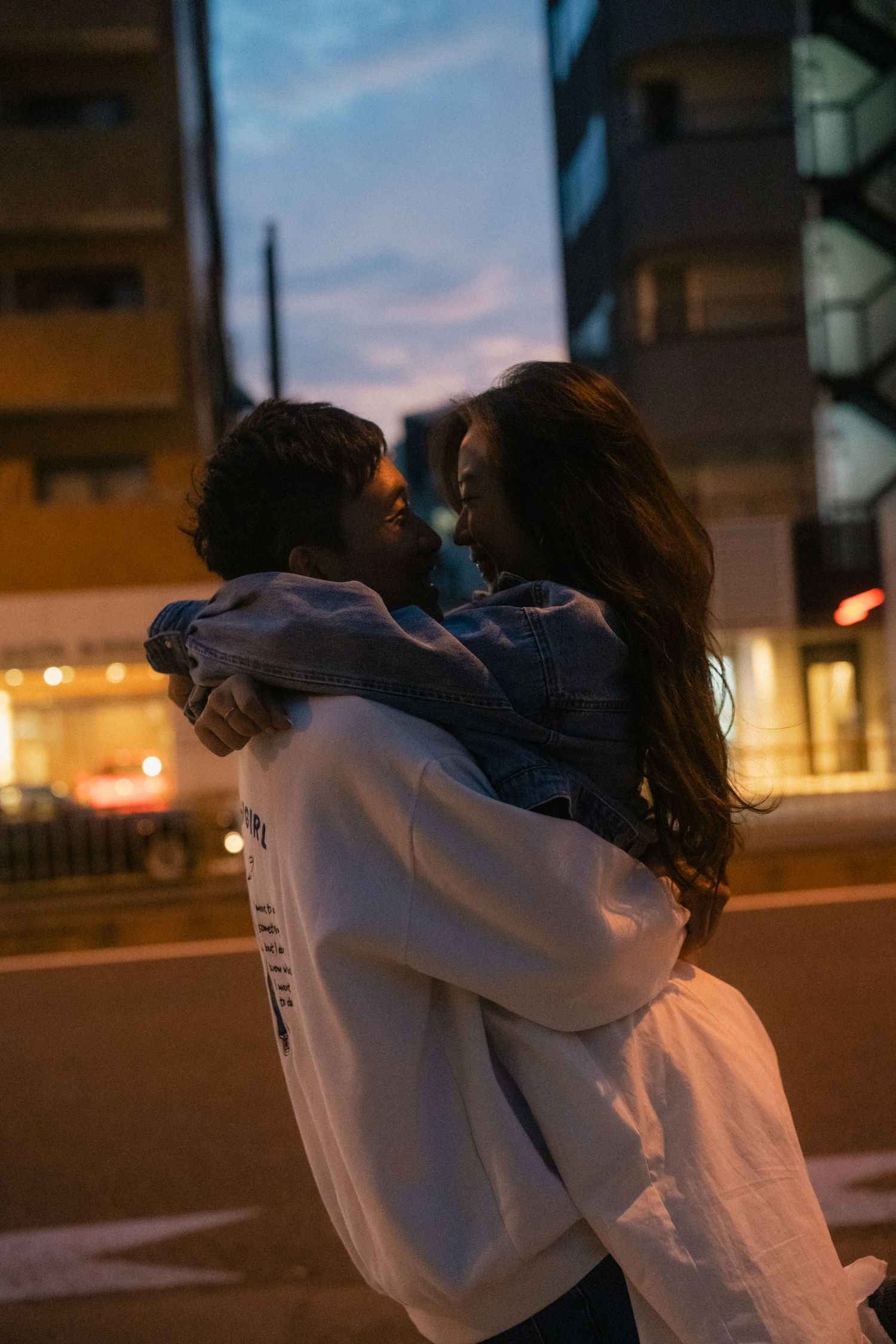 Couple embracing romantically on a city street at dusk, with urban buildings and evening sky in the background.