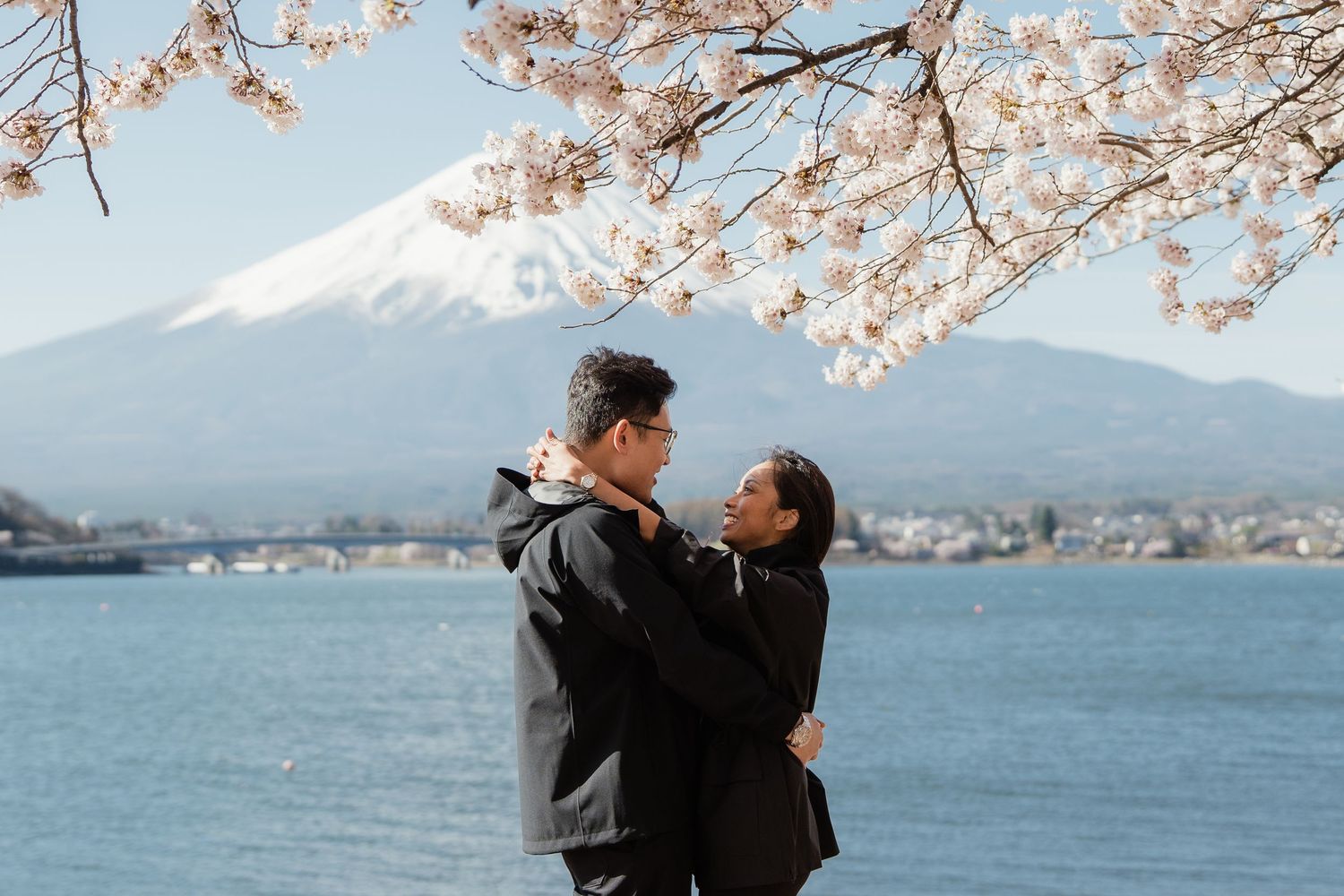 Couple embracing under cherry blossoms with Mount Fuji and lake in background on a clear spring day.