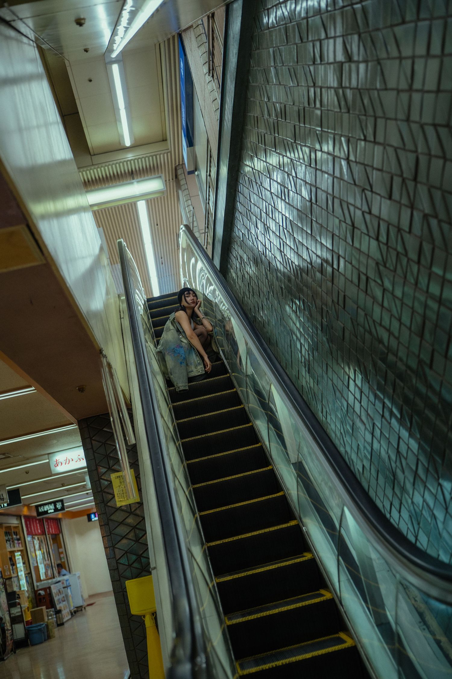 Woman riding an escalator in a modern subway station with tiled walls and bright fluorescent lighting.