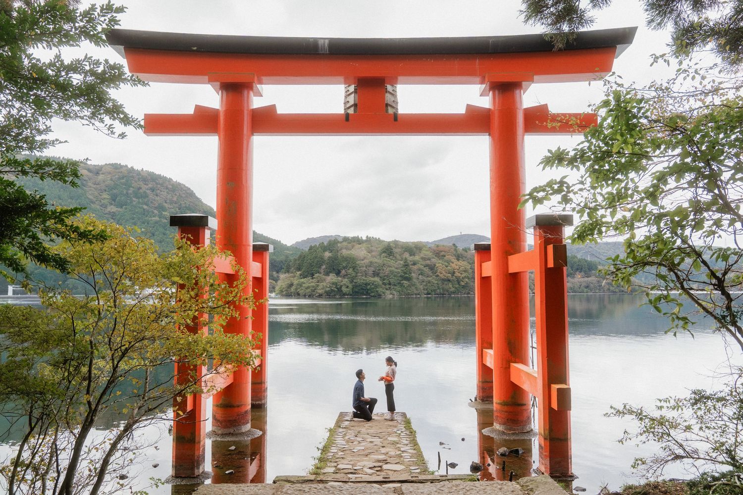 Couple proposing under a red torii gate by a serene lake in Hakone, Japan, surrounded by lush green mountains.