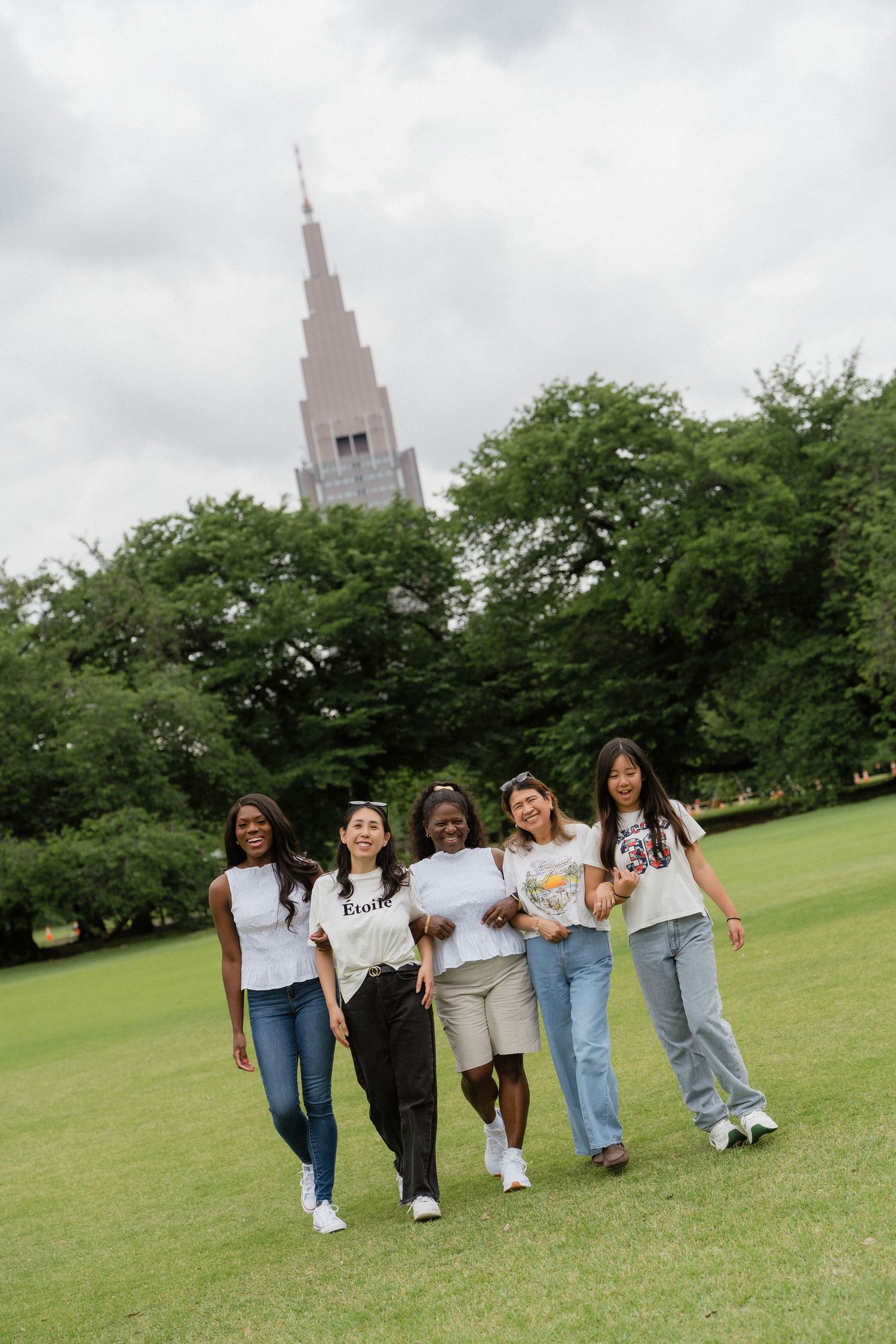 Five diverse young women walking together on green grass with trees and a tall spire building in the background.