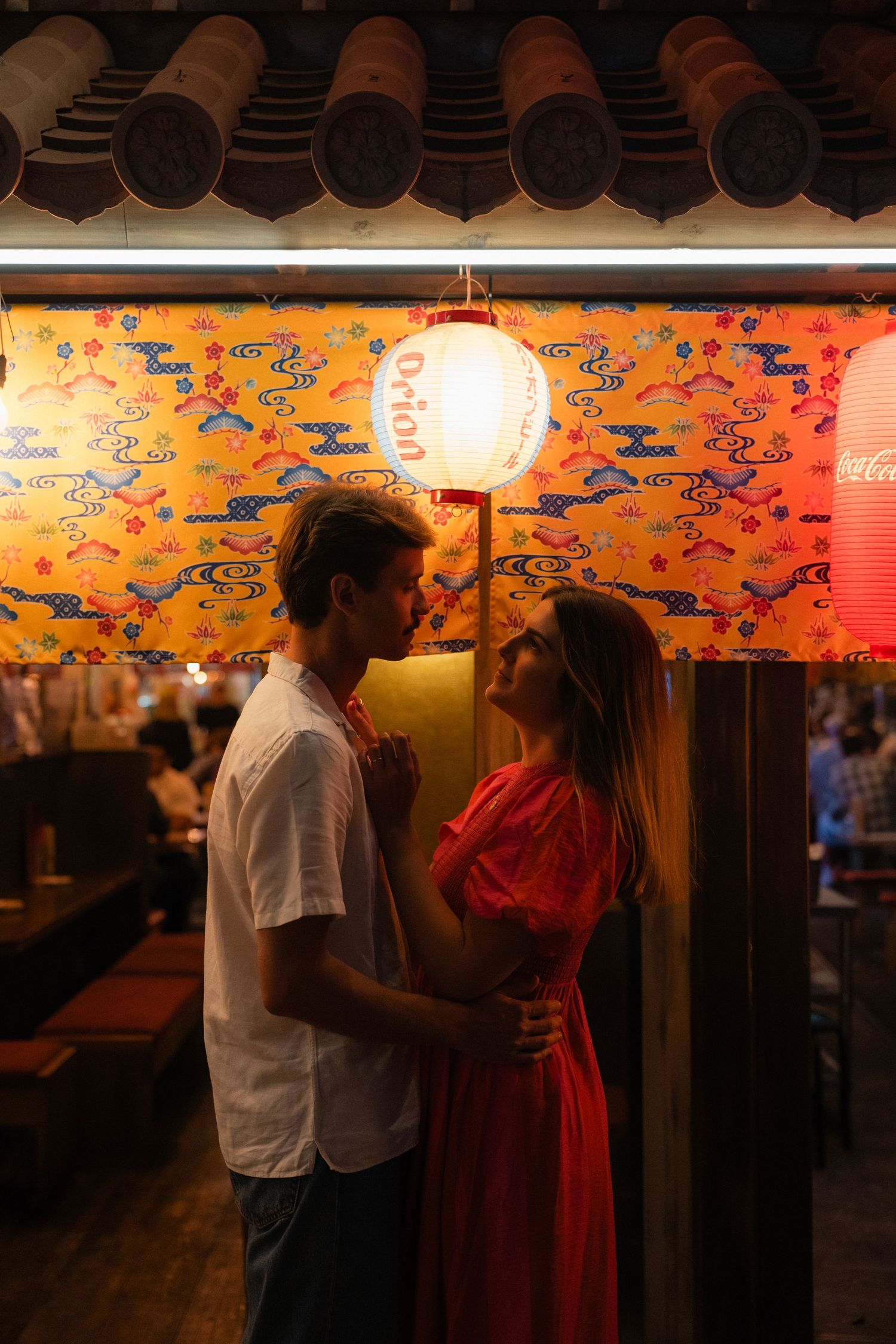 Romantic couple embracing under a glowing paper lantern outside a Japanese restaurant with colorful decor.