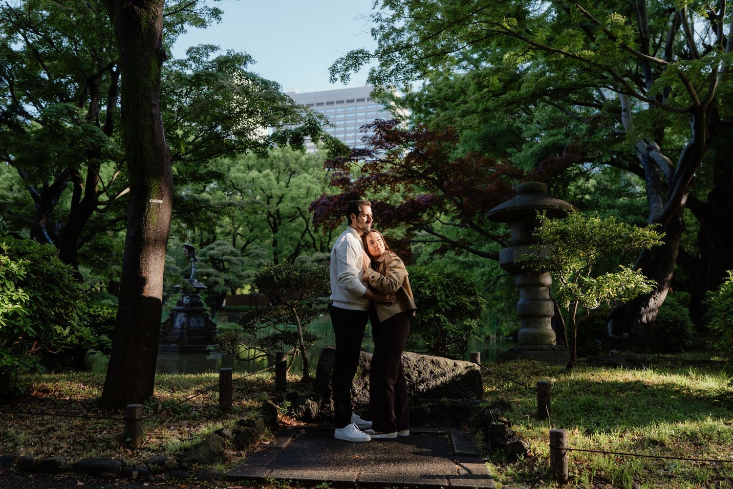 Couple embracing in a lush Japanese garden with stone lanterns and green trees in soft natural light.
