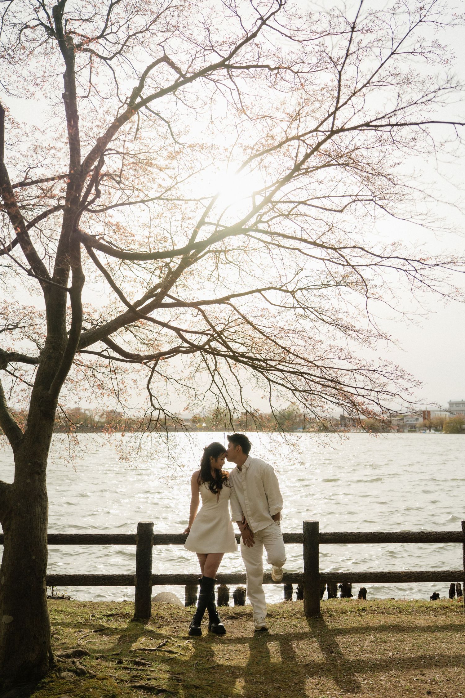 Couple sharing a romantic moment under cherry blossom tree by a serene lake, dressed in white for an engagement photo.