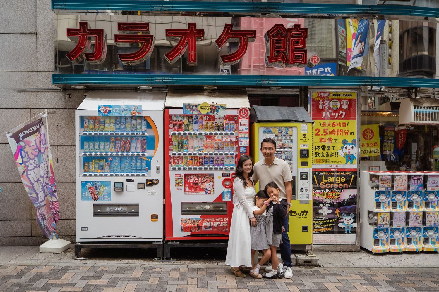 Family posing in front of colorful vending machines outside a karaoke building in Japan.