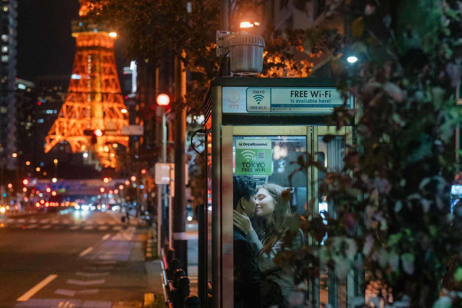 Woman using free Wi-Fi phone booth at night with Tokyo Tower illuminated in background on busy city street.