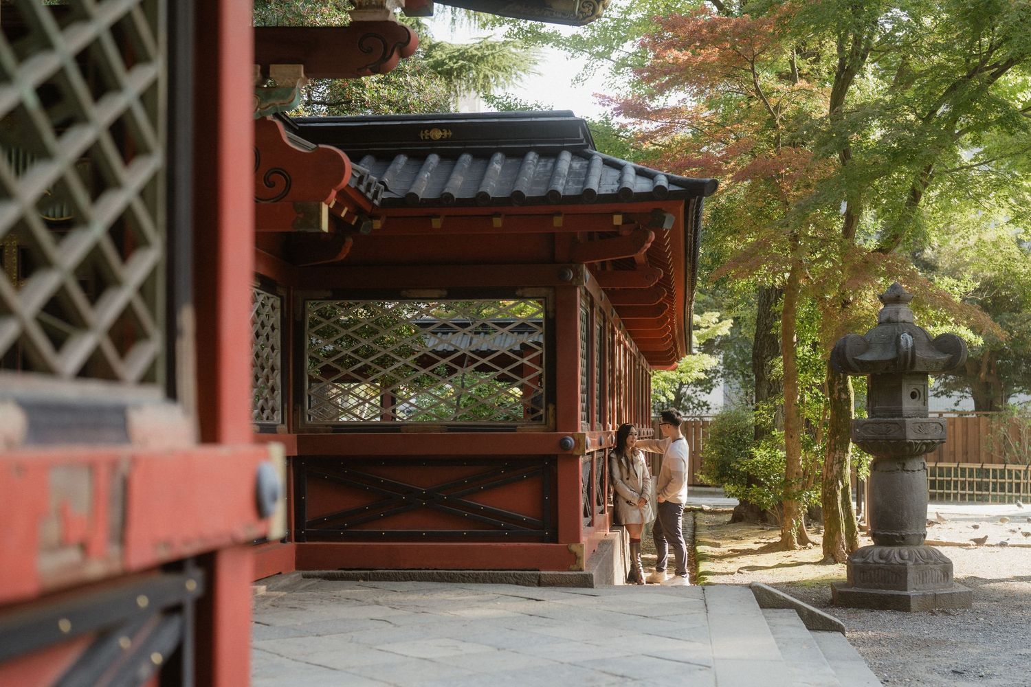 Red wooden Japanese temple corridor with stone lantern and lush green trees in a serene traditional shrine setting.