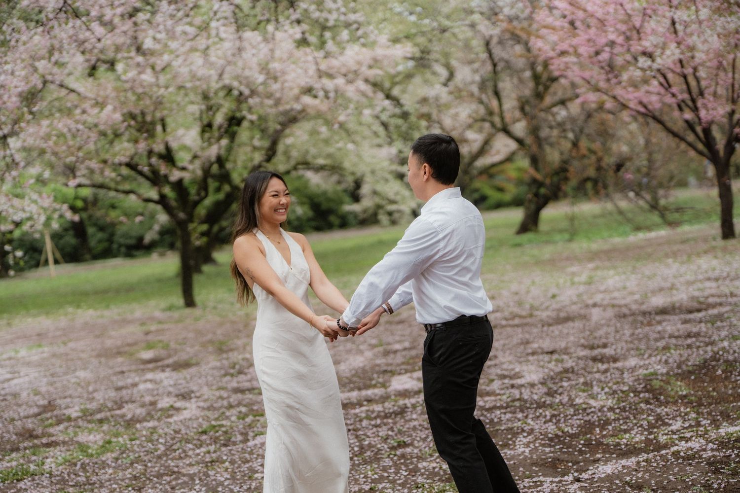 Couple dancing among blooming cherry blossom trees, woman in white gown holding hands with man in white shirt and black pants.