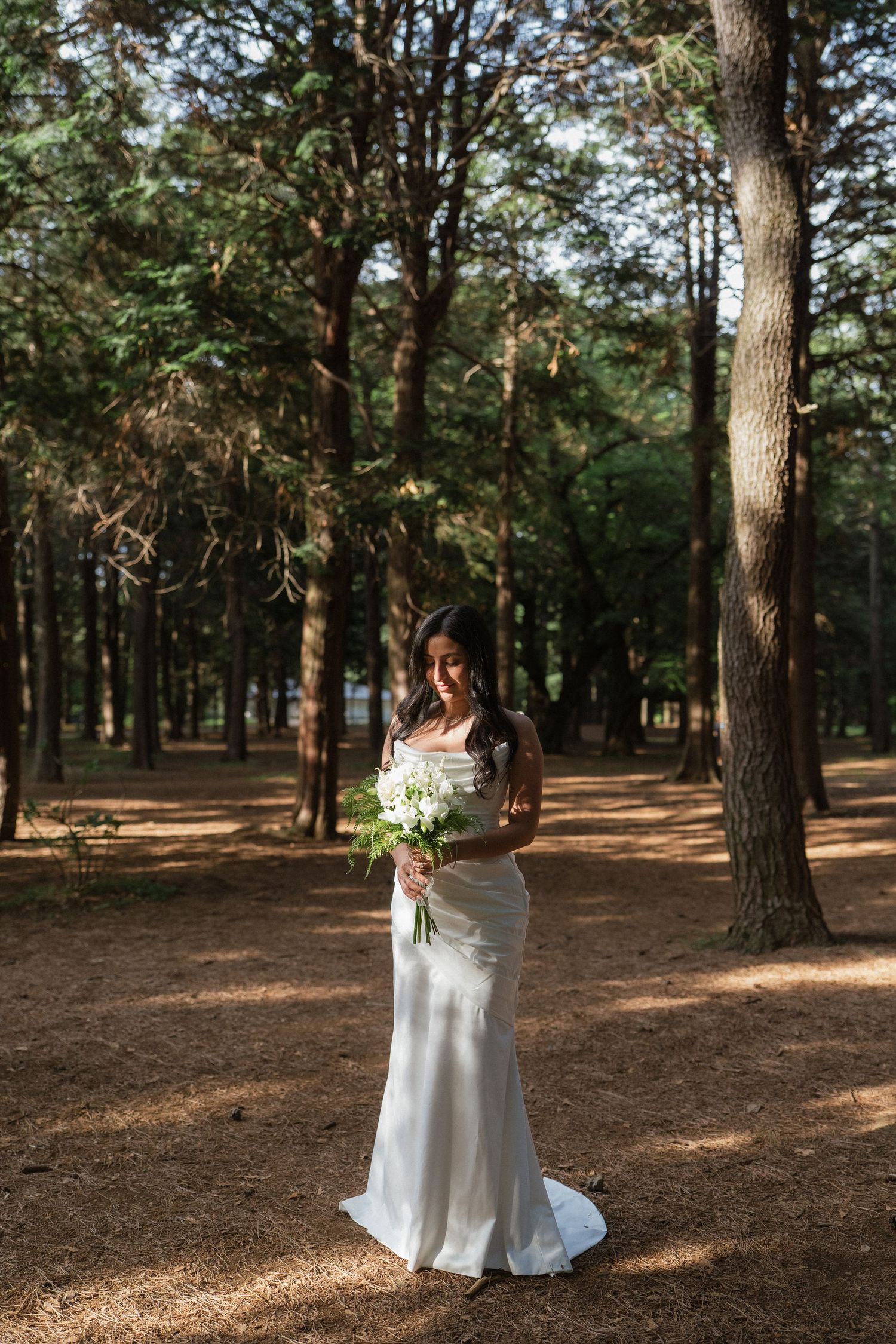Bride in white fitted gown holding white bouquet standing among tall pine trees in dappled sunlight.