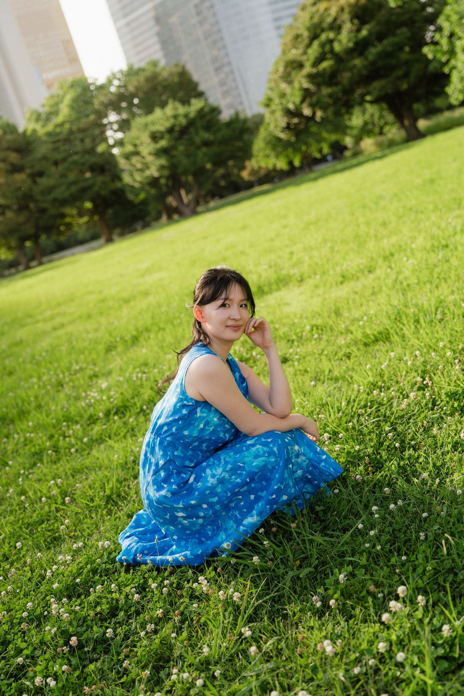 Young woman in blue floral dress sitting on green grass in a park with trees and city buildings in background.