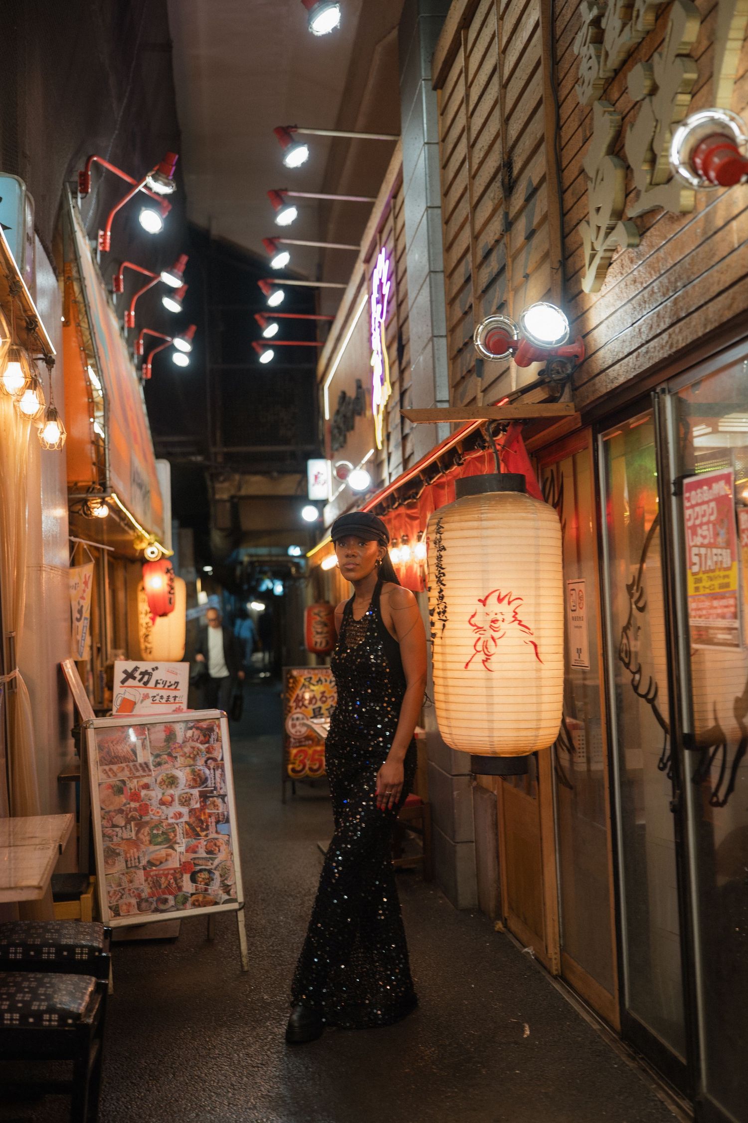 Woman in black sequin gown poses in a vibrant Japanese alley at night, surrounded by glowing lanterns and restaurant signs.