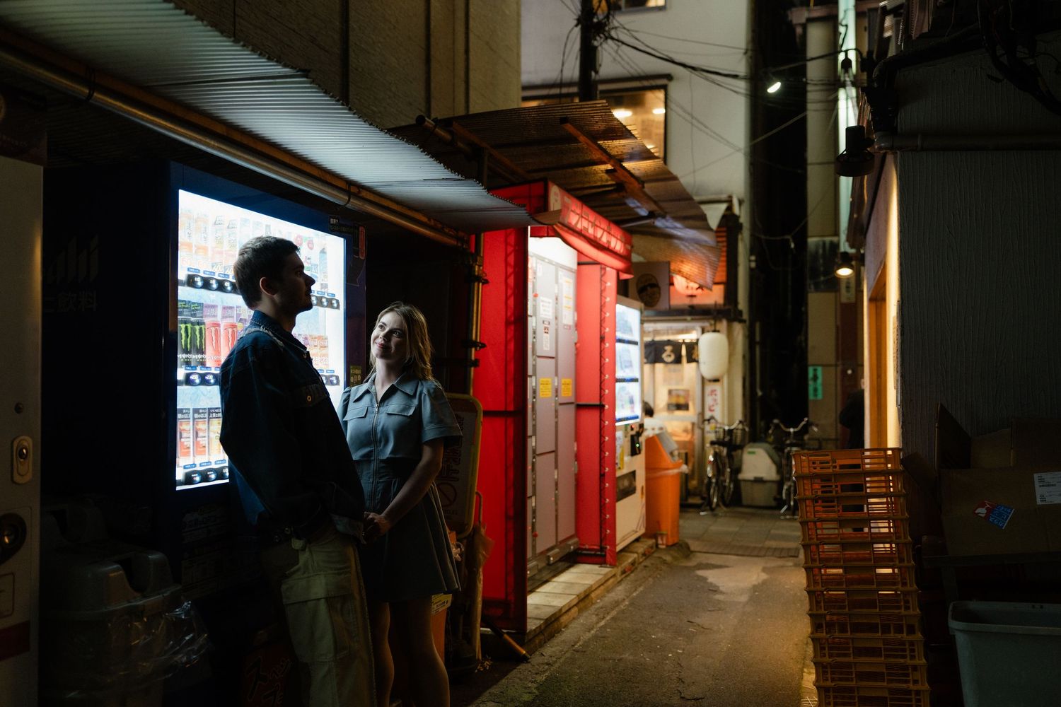 Two people stand in a dimly lit alley beside a glowing red door and illuminated vending machine at night.