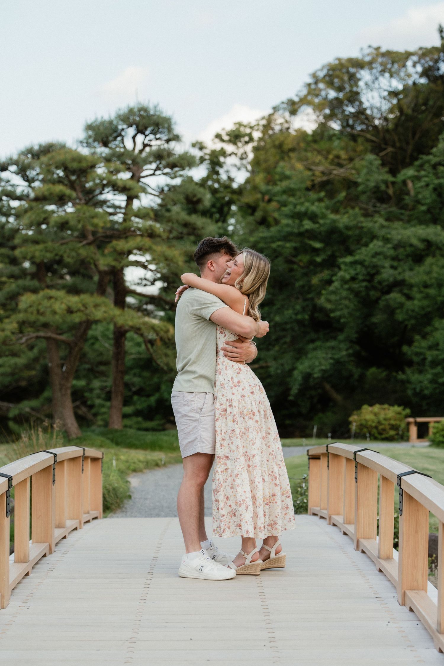 Couple embracing and kissing on a wooden bridge surrounded by lush green trees in a serene park setting.