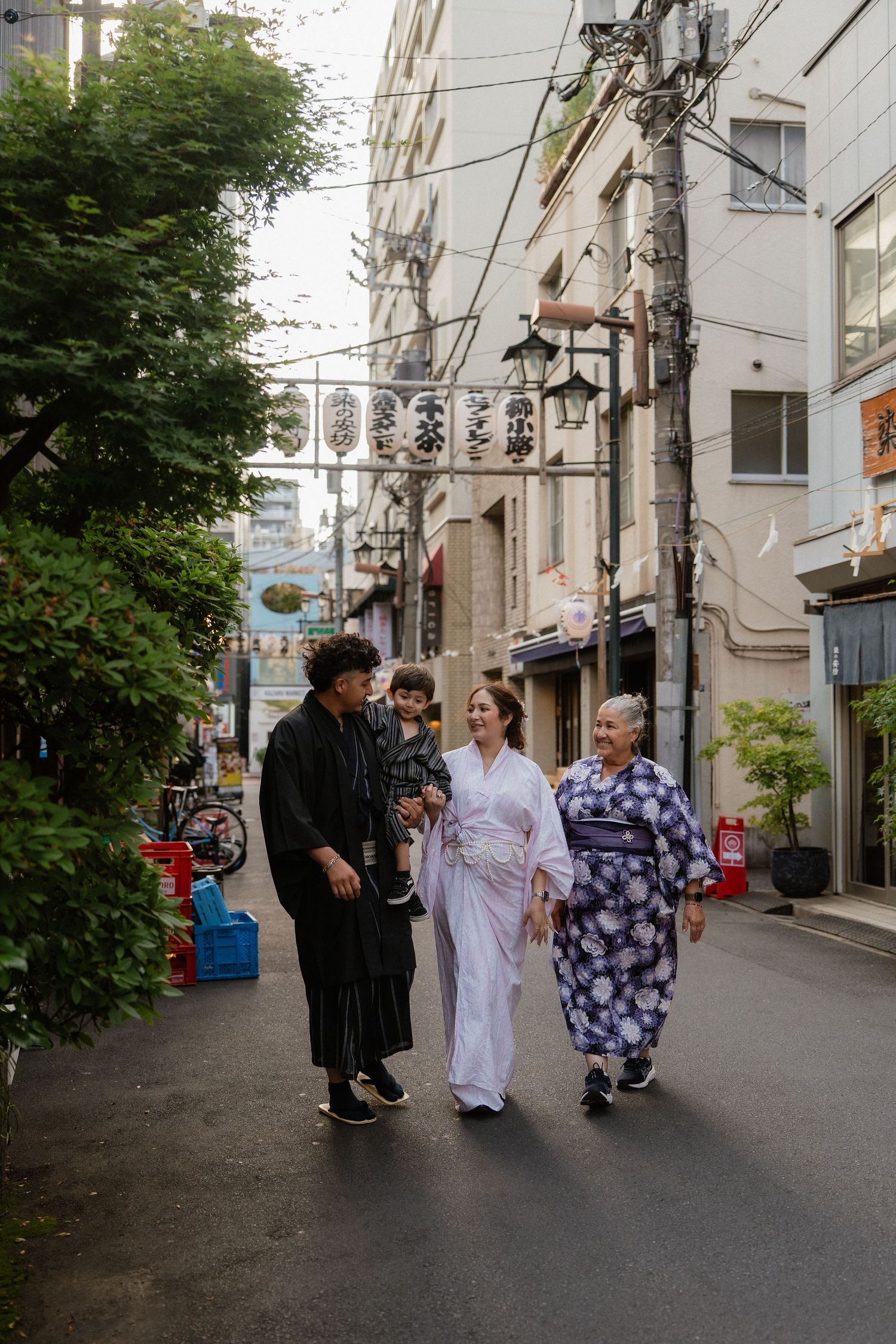 Family strolling a Japanese alley in traditional kimonos and yukata, surrounded by urban buildings and lush greenery.