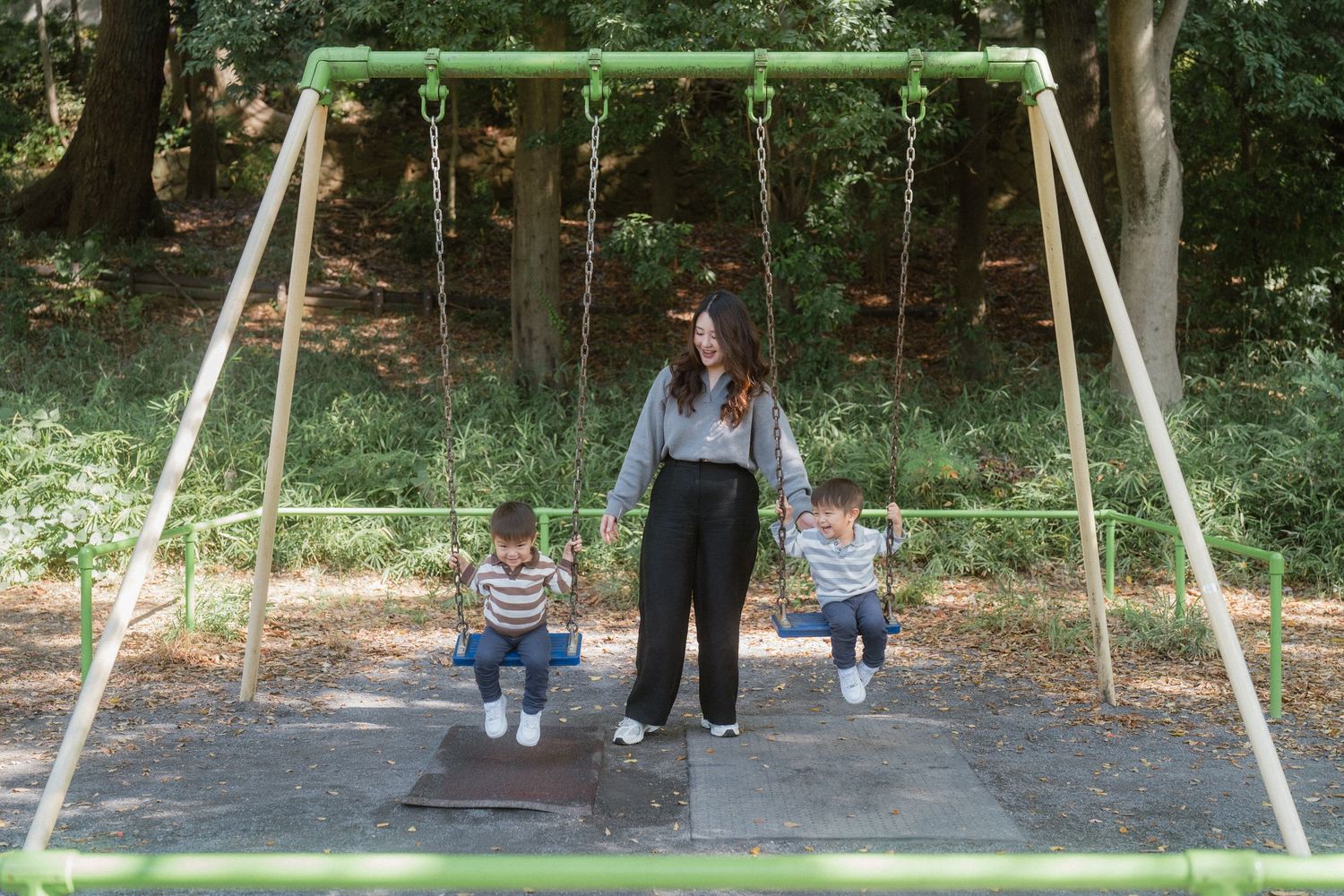 A mother pushes two young children on a green swing set in a shaded outdoor playground.