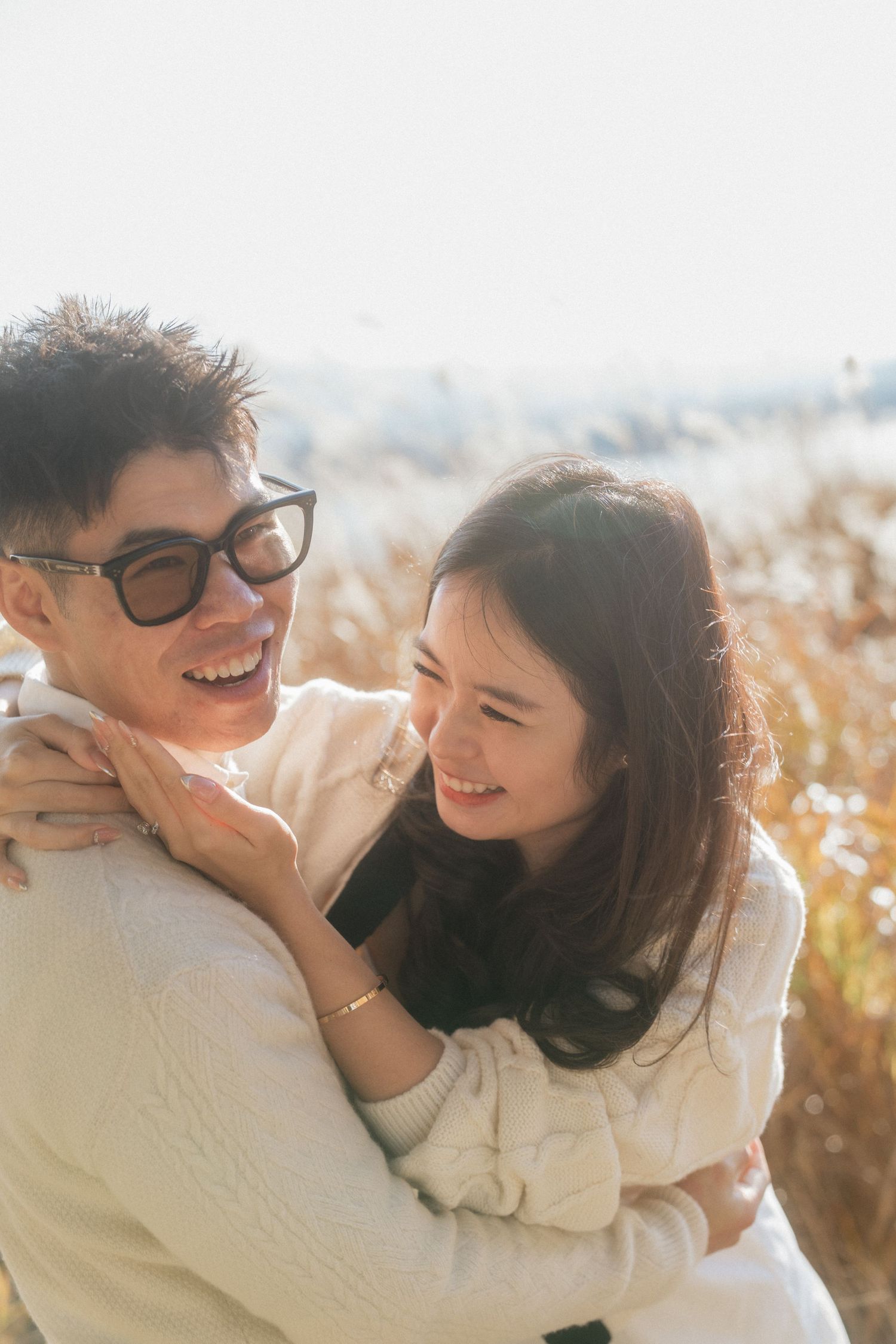 Happy Asian couple embracing outdoors in a sunny field, wearing matching white sweaters, smiling joyfully at each other.