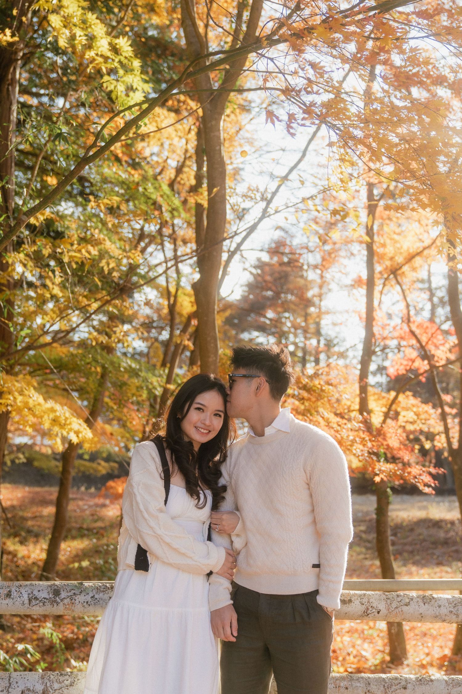 Couple embracing in autumn park, woman in white dress smiling, man in cream sweater, colorful fall foliage background.