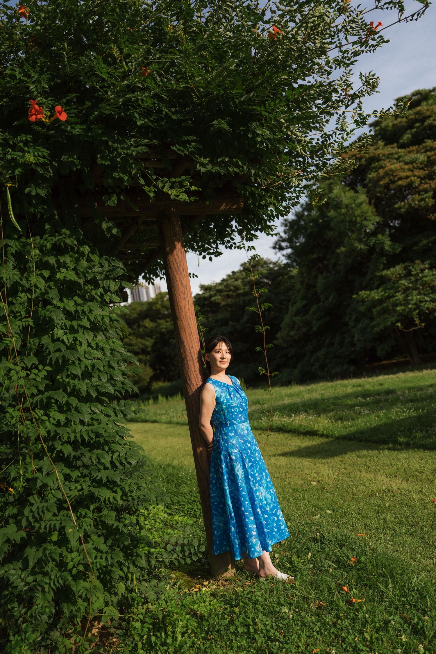 A young woman in a blue floral midi dress stands gracefully beside a tree trunk in a lush green park.