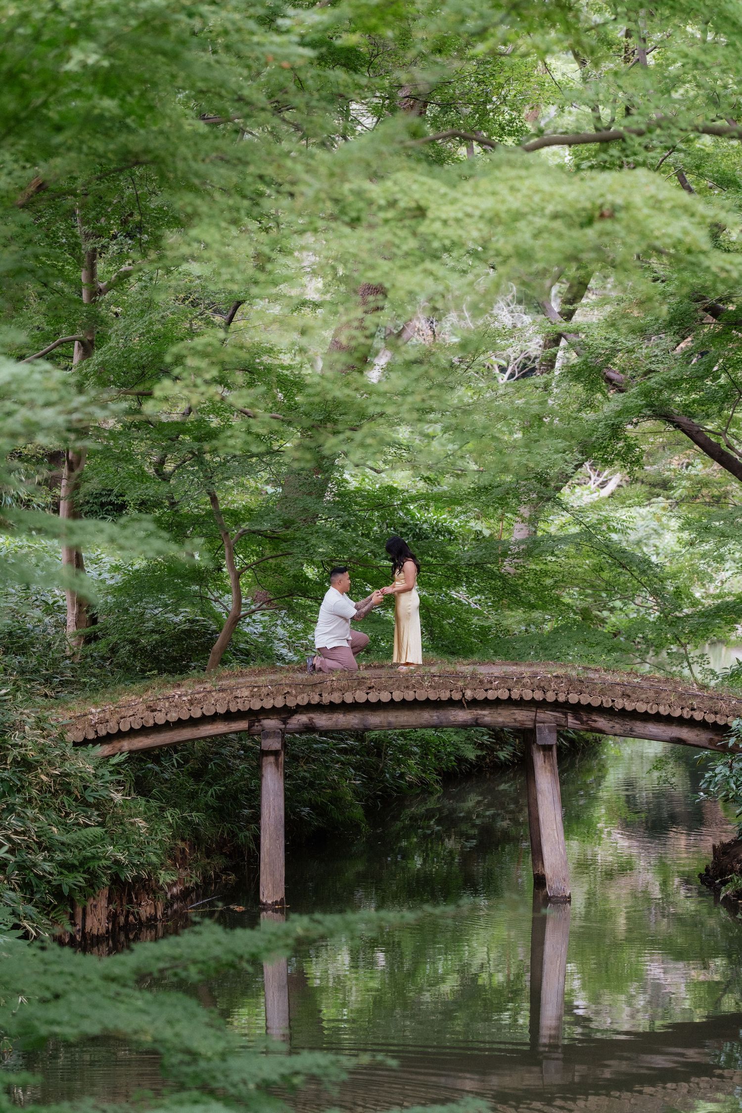 Couple sharing a romantic moment on an arched wooden bridge over a tranquil pond in a lush green Japanese garden.