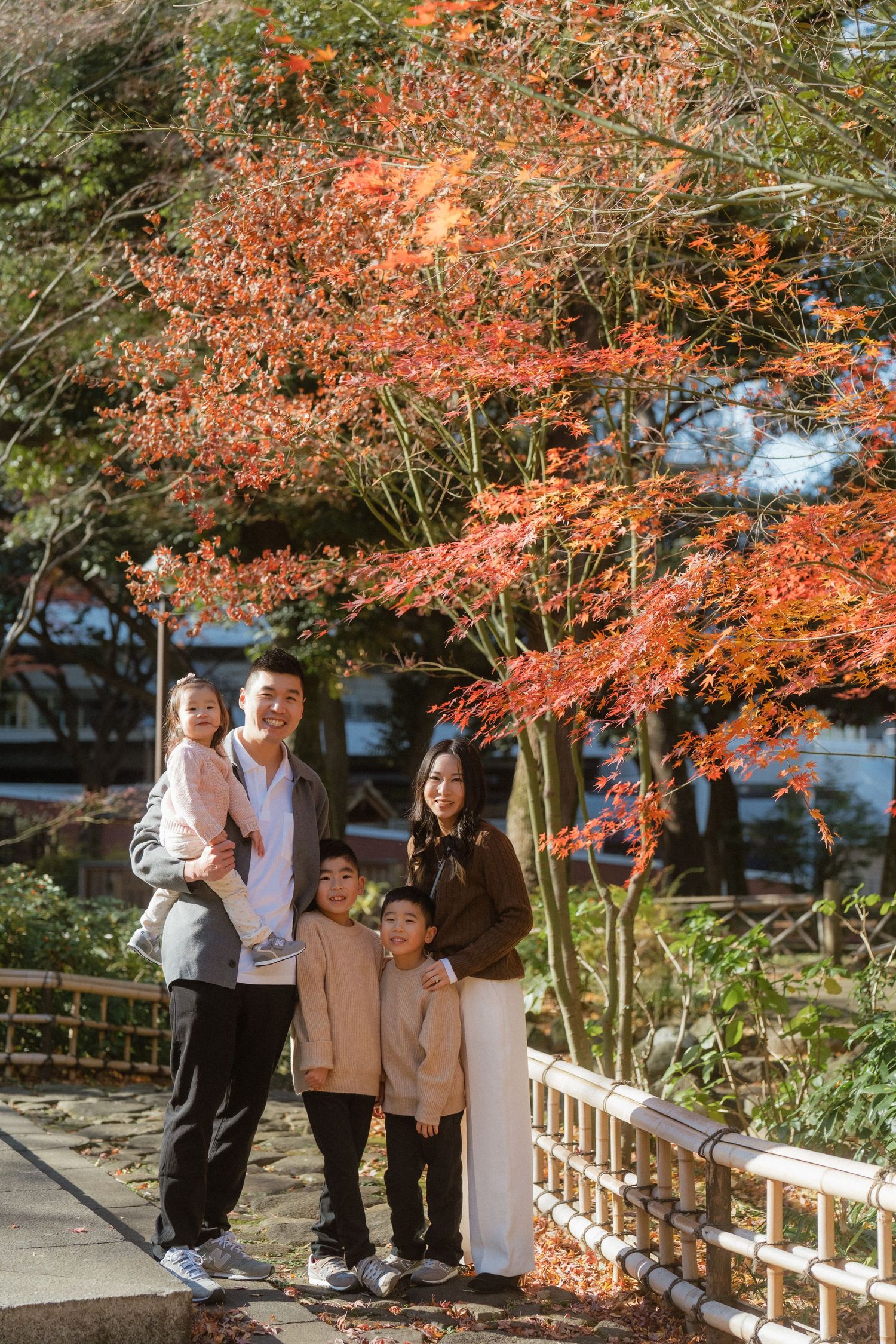 Family of four posing under vibrant red autumn maple tree by a white fence in a Japanese garden.