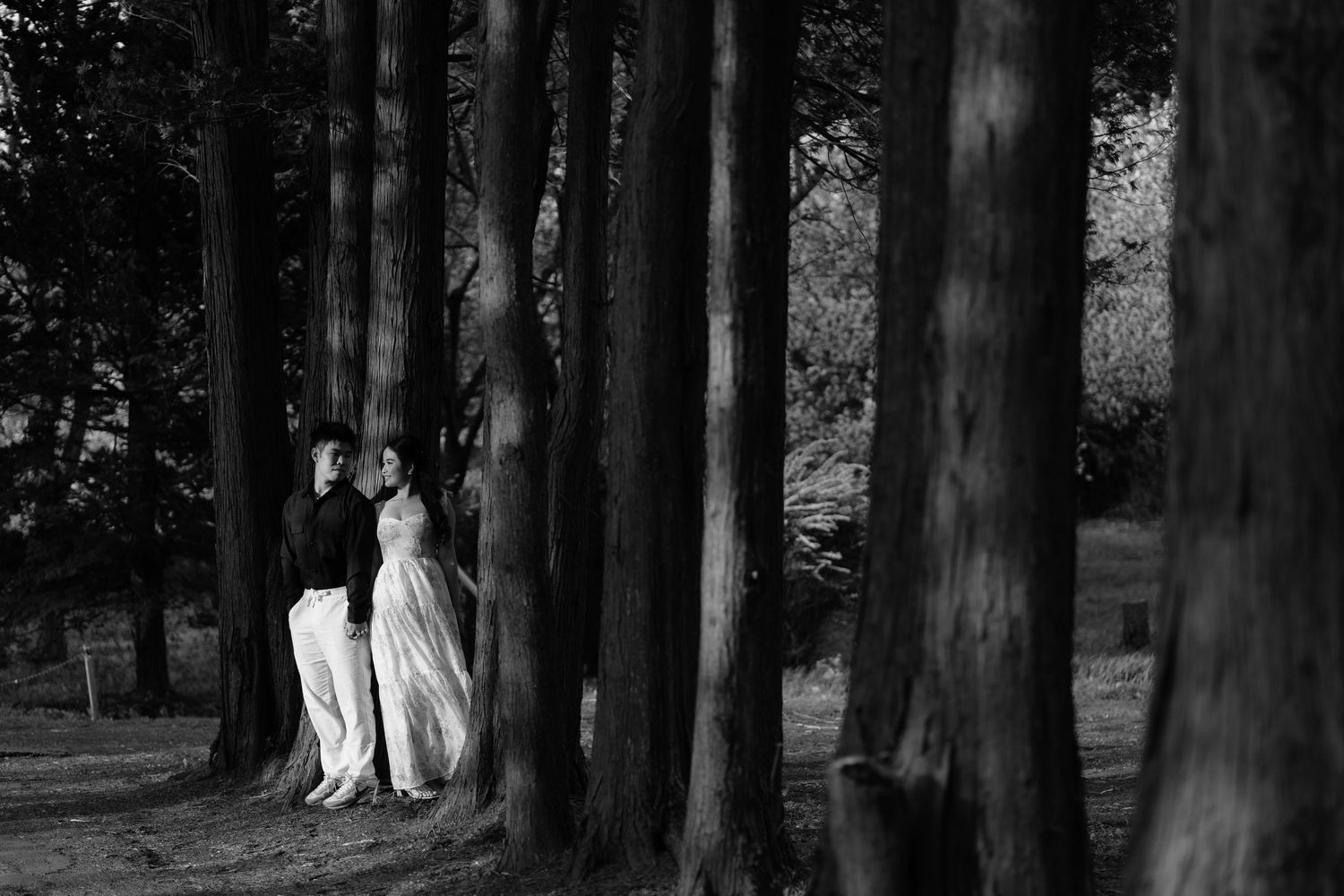 Black and white photo of a couple in wedding attire standing among tall trees in a forest.