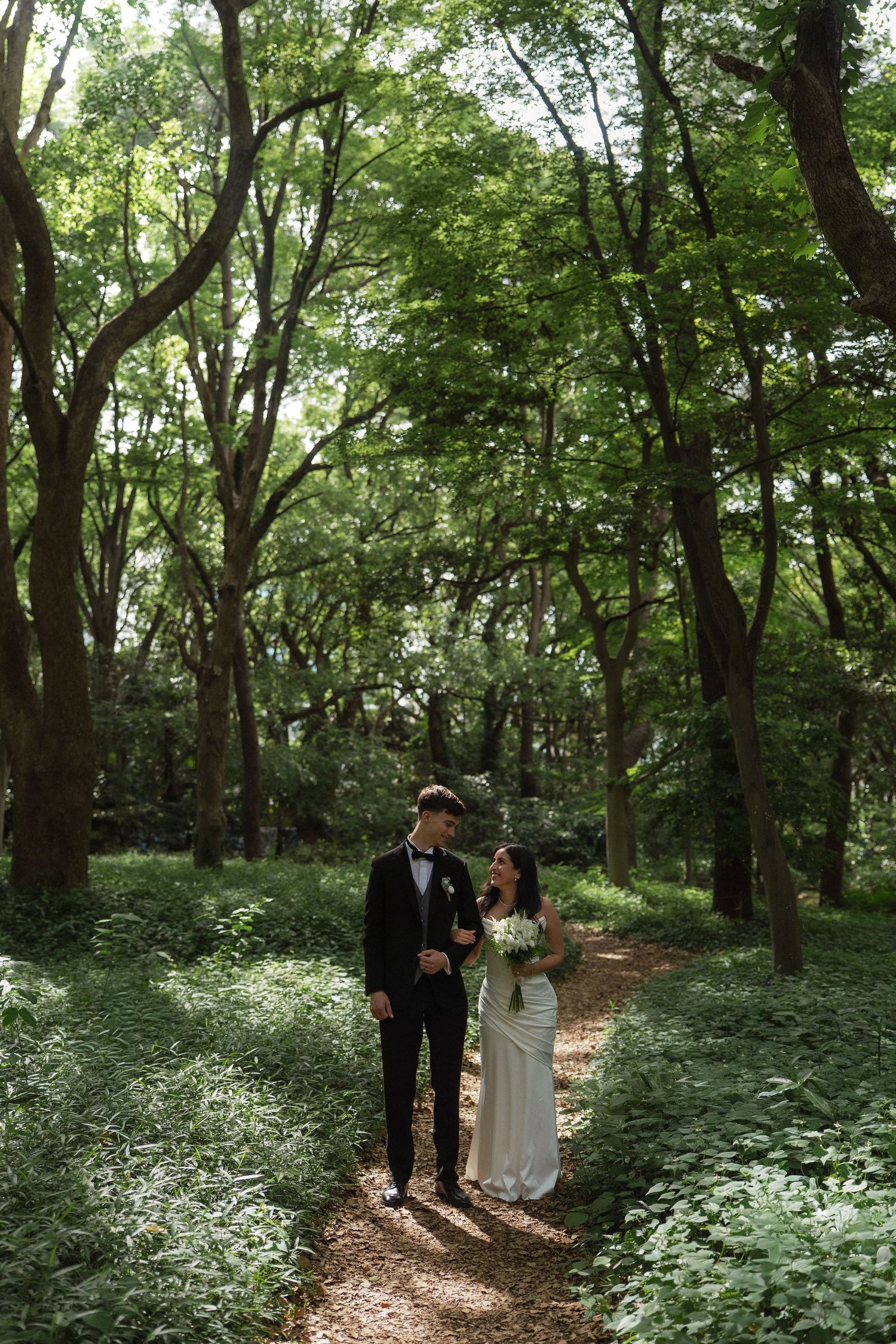 Bride and groom walk hand in hand along a forest path surrounded by tall trees.