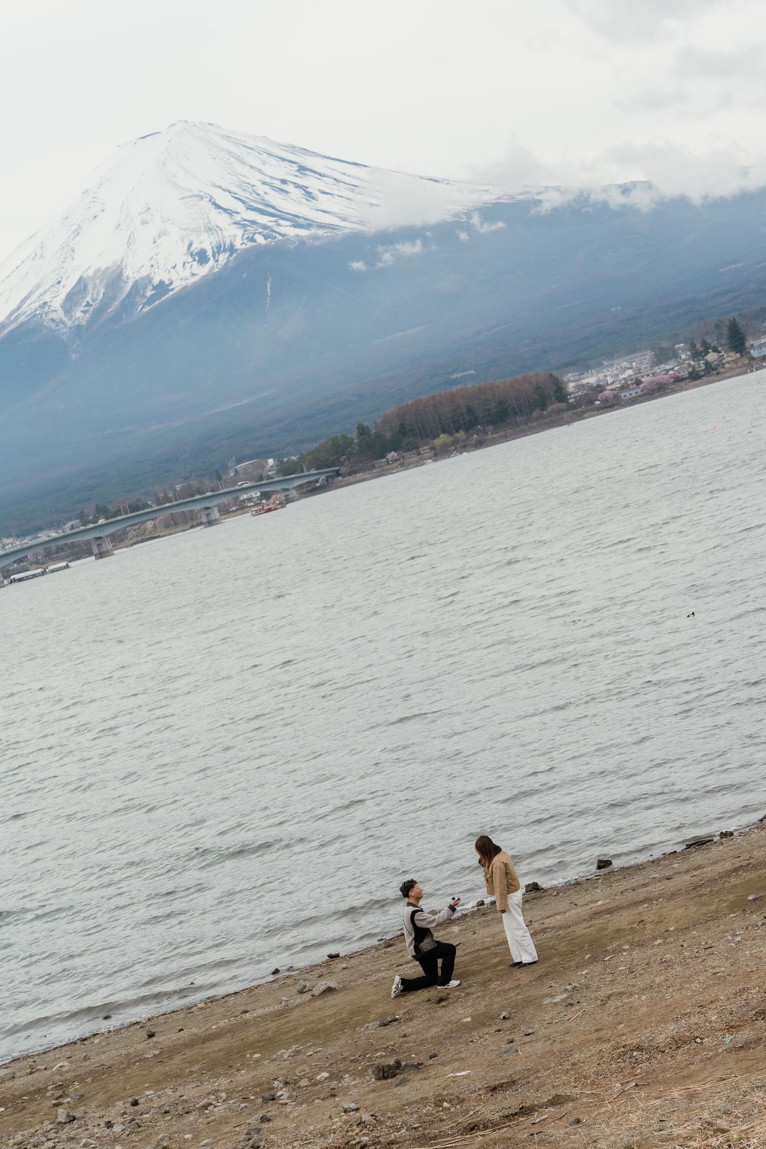 Man proposes on bended knee to woman by Lake Kawaguchiko with snow-capped Mount Fuji in background.