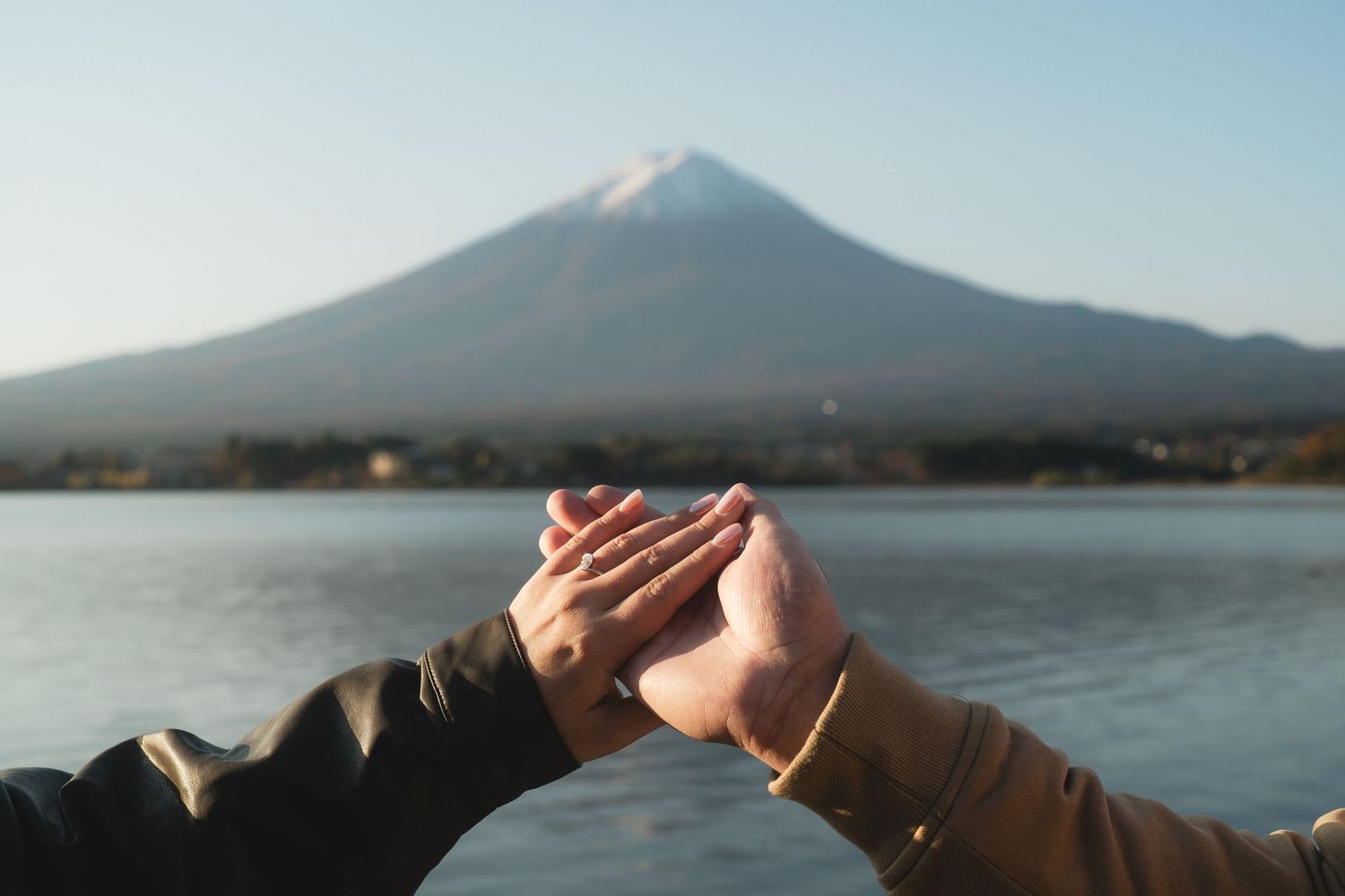 Couple holding hands near Mount Fuji with serene lake and snow-capped peak in background.