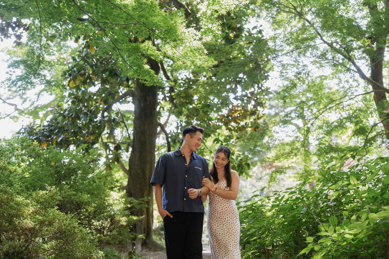 Couple walking together on a garden path surrounded by lush green trees in a serene outdoor setting.