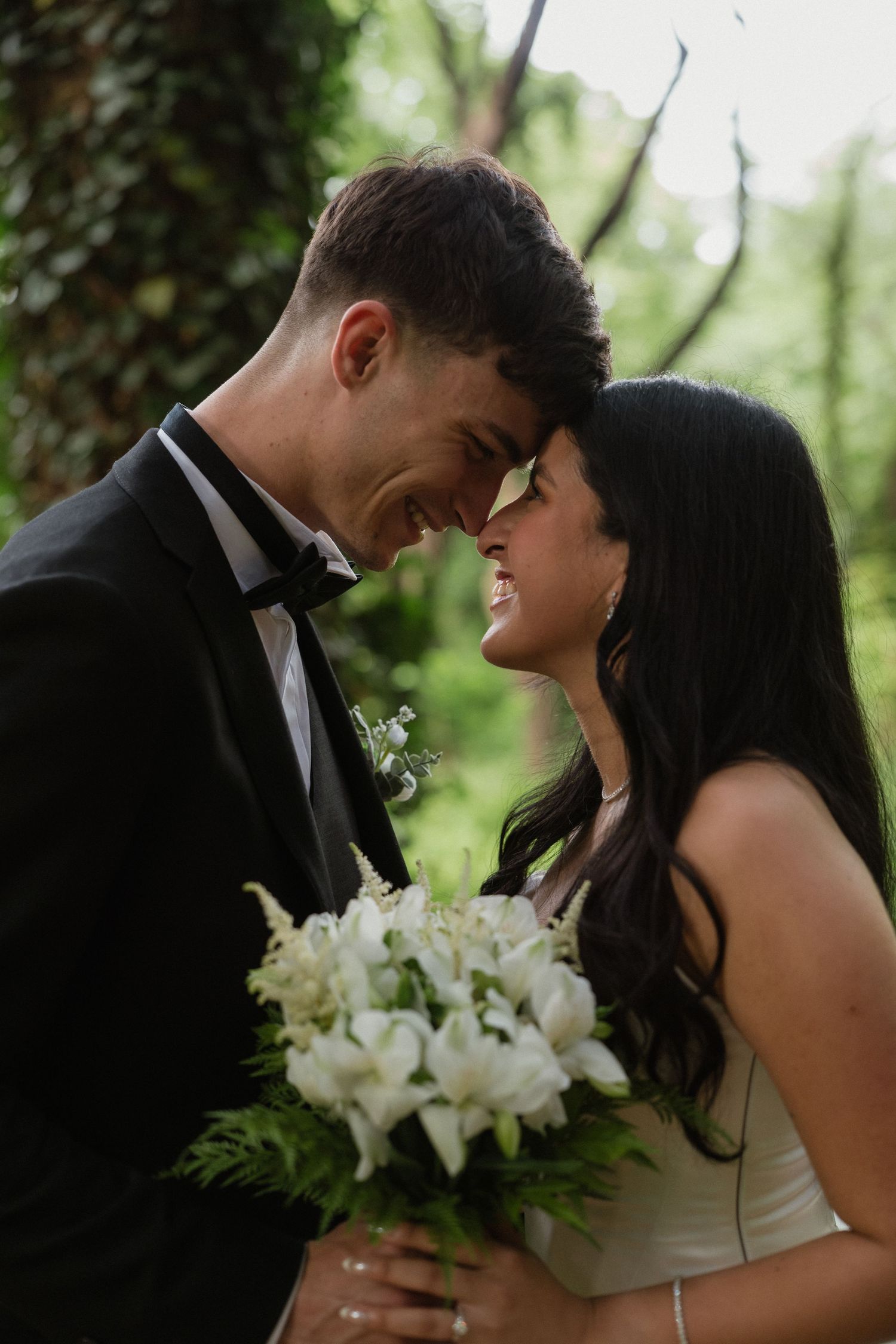 A smiling bride and groom touch foreheads while the bride holds a white lily bouquet in a lush green outdoor setting.