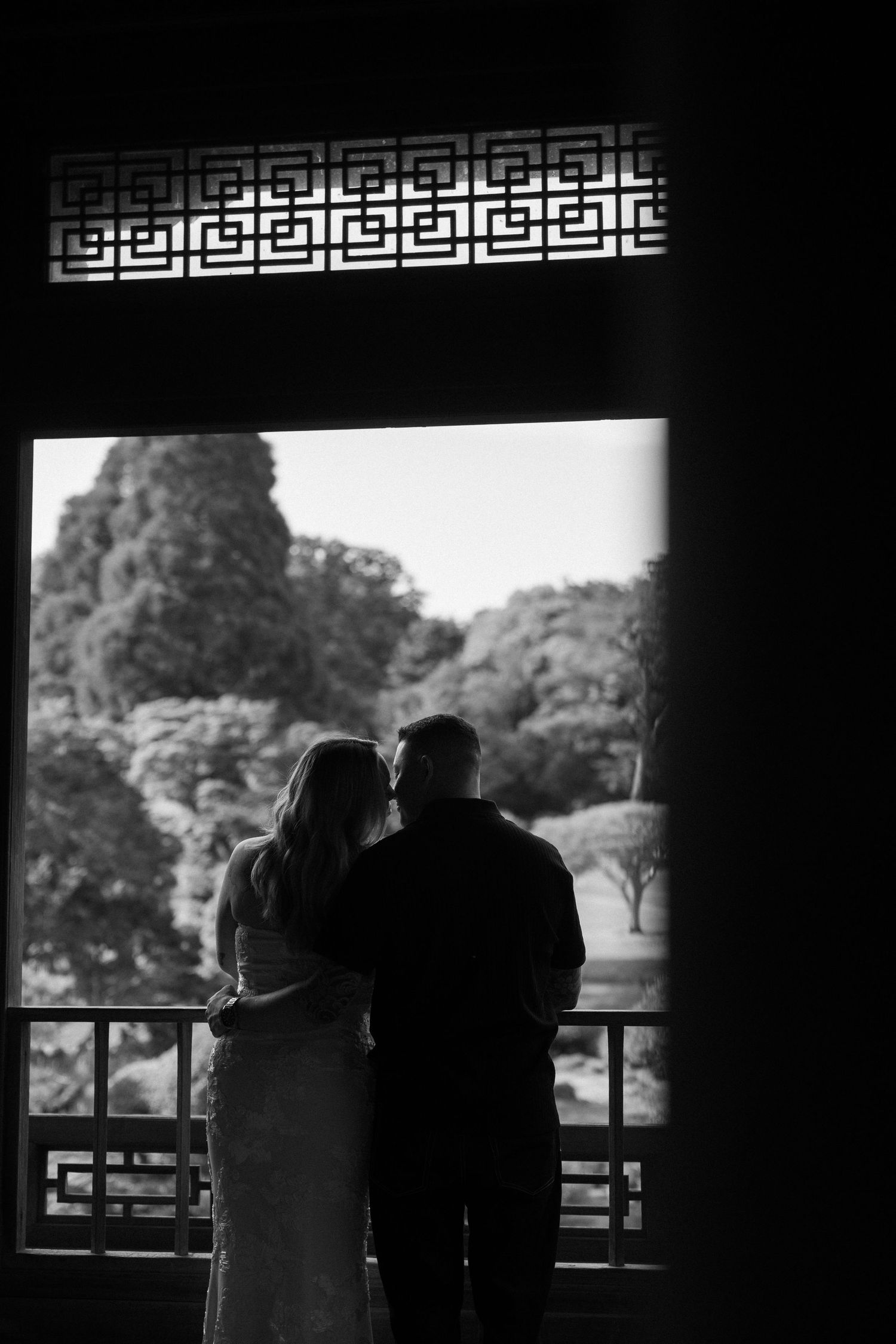 Black and white silhouette of a couple embracing in a Chinese garden pavilion doorway with lush trees in the background.