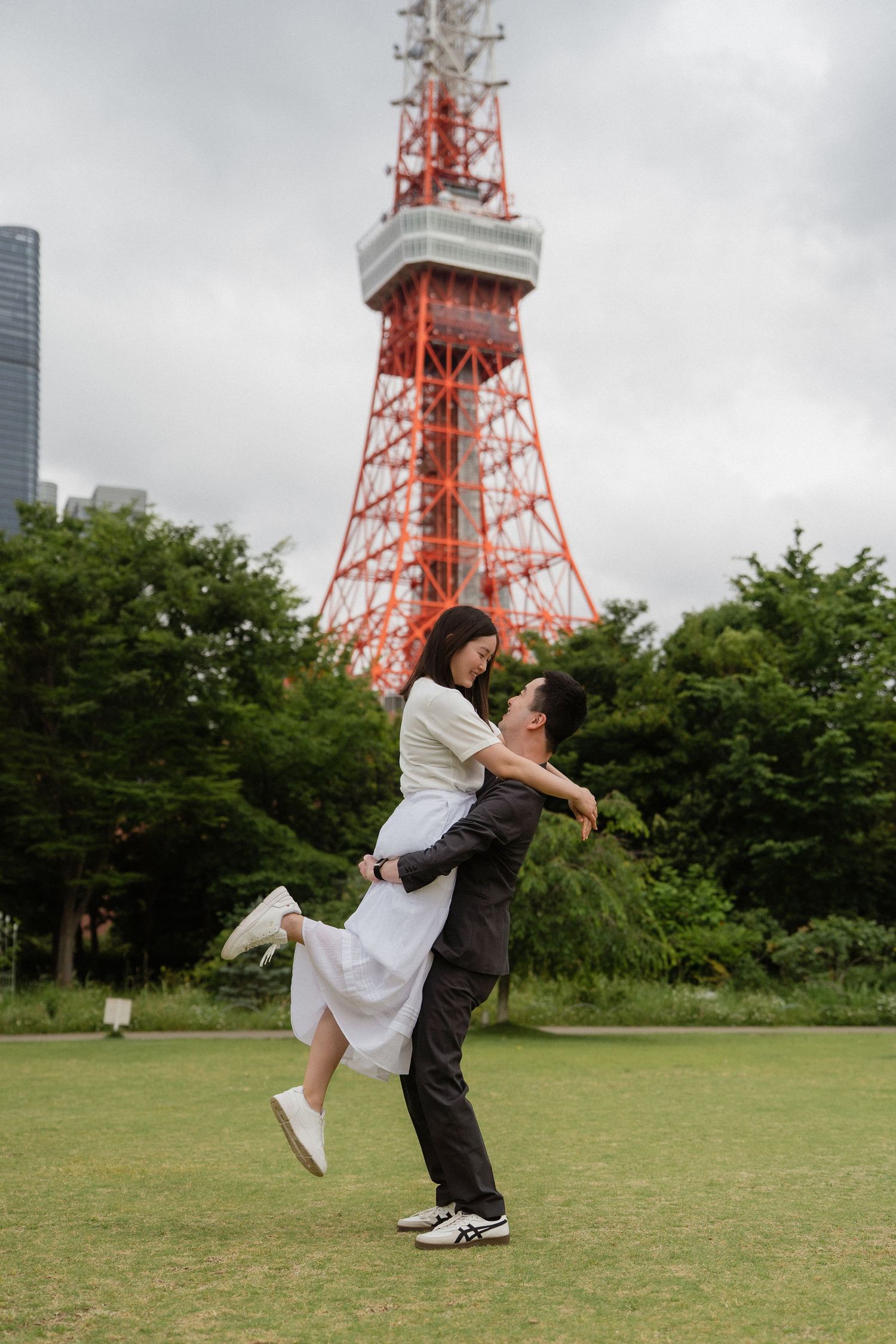 Couple embracing in front of Tokyo Tower on a green lawn, man lifting woman in white dress, romantic outdoor photo.