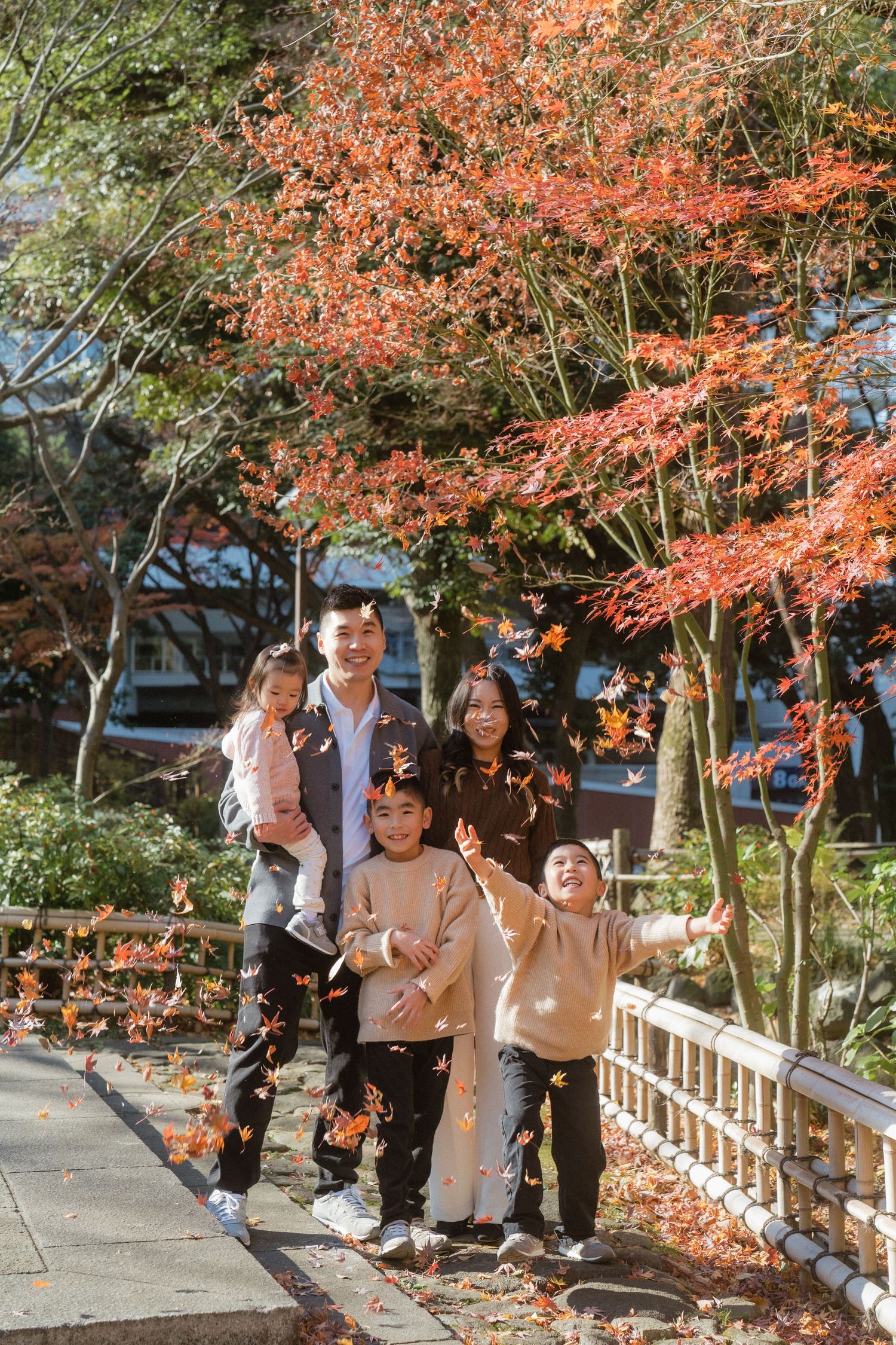 Family of five joyfully tossing autumn leaves under vibrant red maple trees in a scenic park setting.