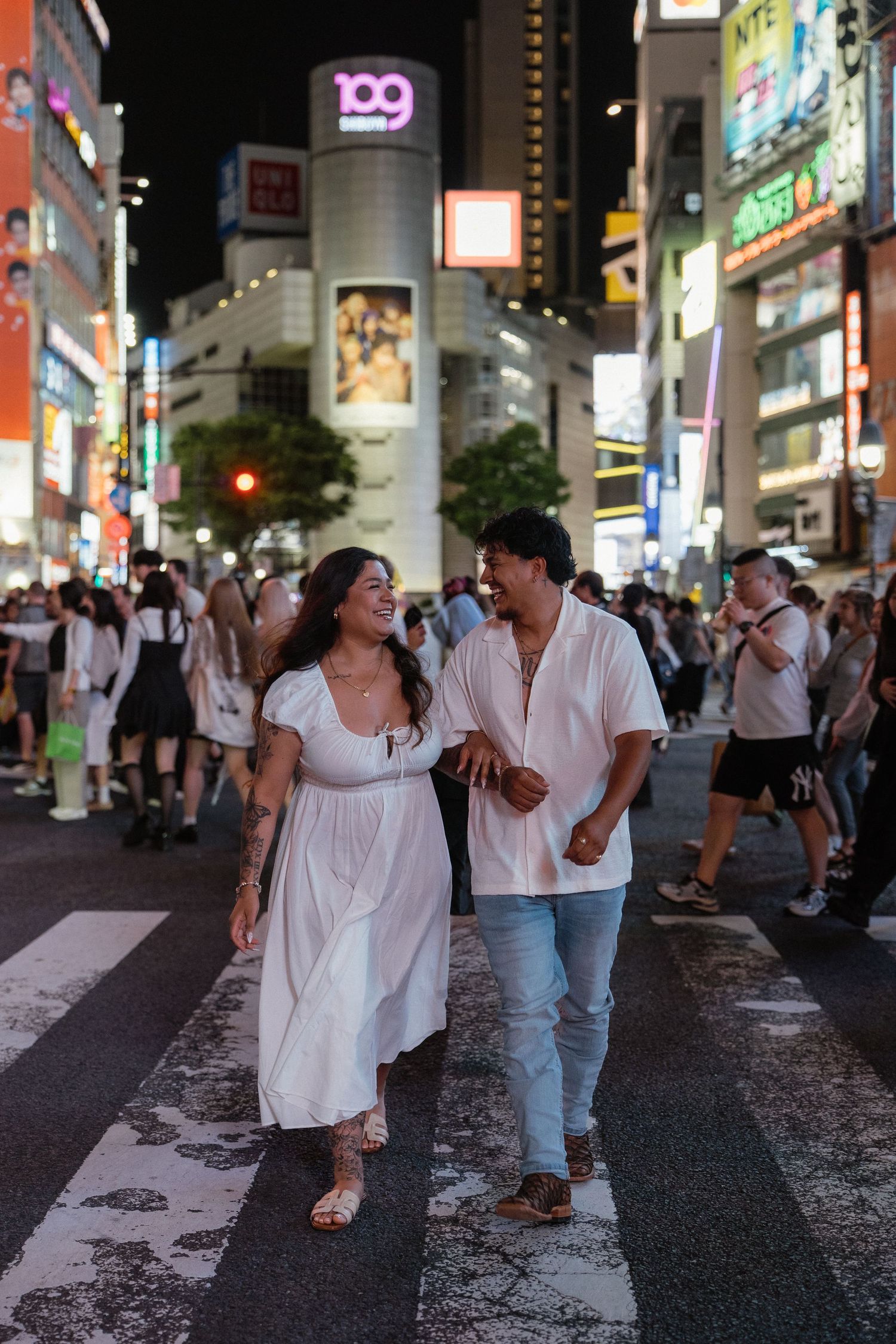 Couple in white outfits laughing while crossing Shibuya Crossing at night in Tokyo, Japan.