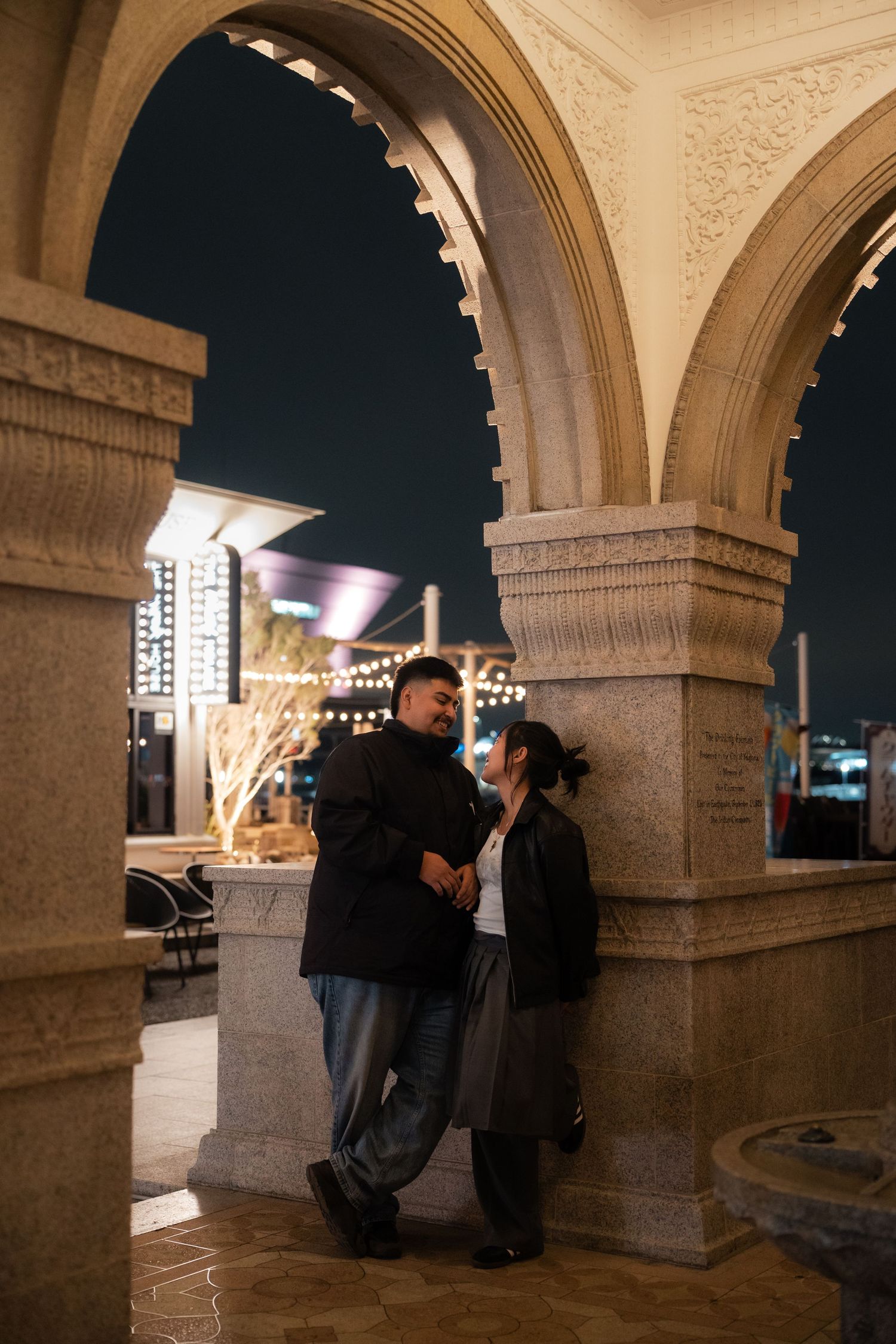 Couple sharing a romantic moment under stone arches at night, with string lights glowing in the background.