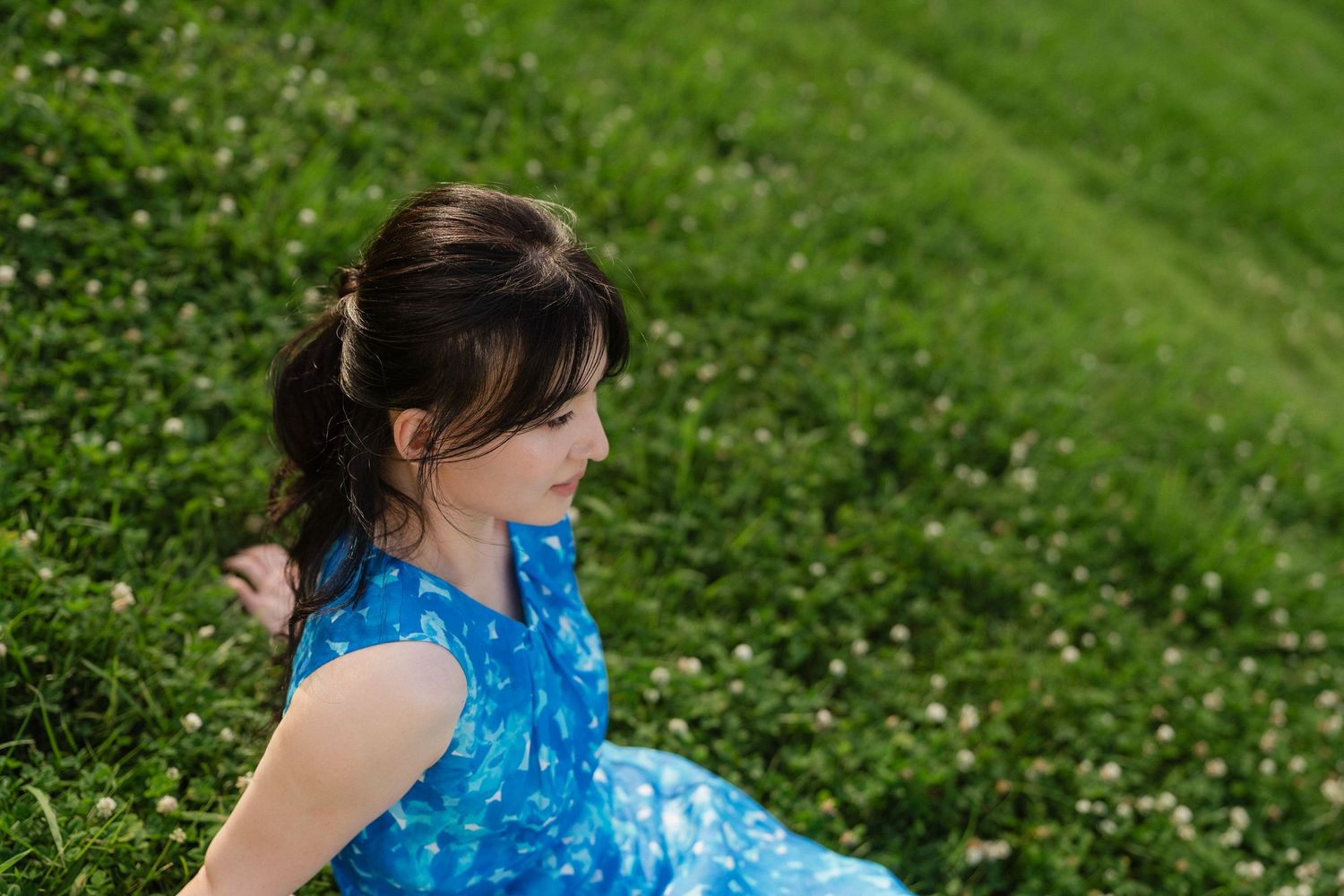 Young girl in blue floral dress sitting on lush green grass, looking away in a peaceful outdoor setting.